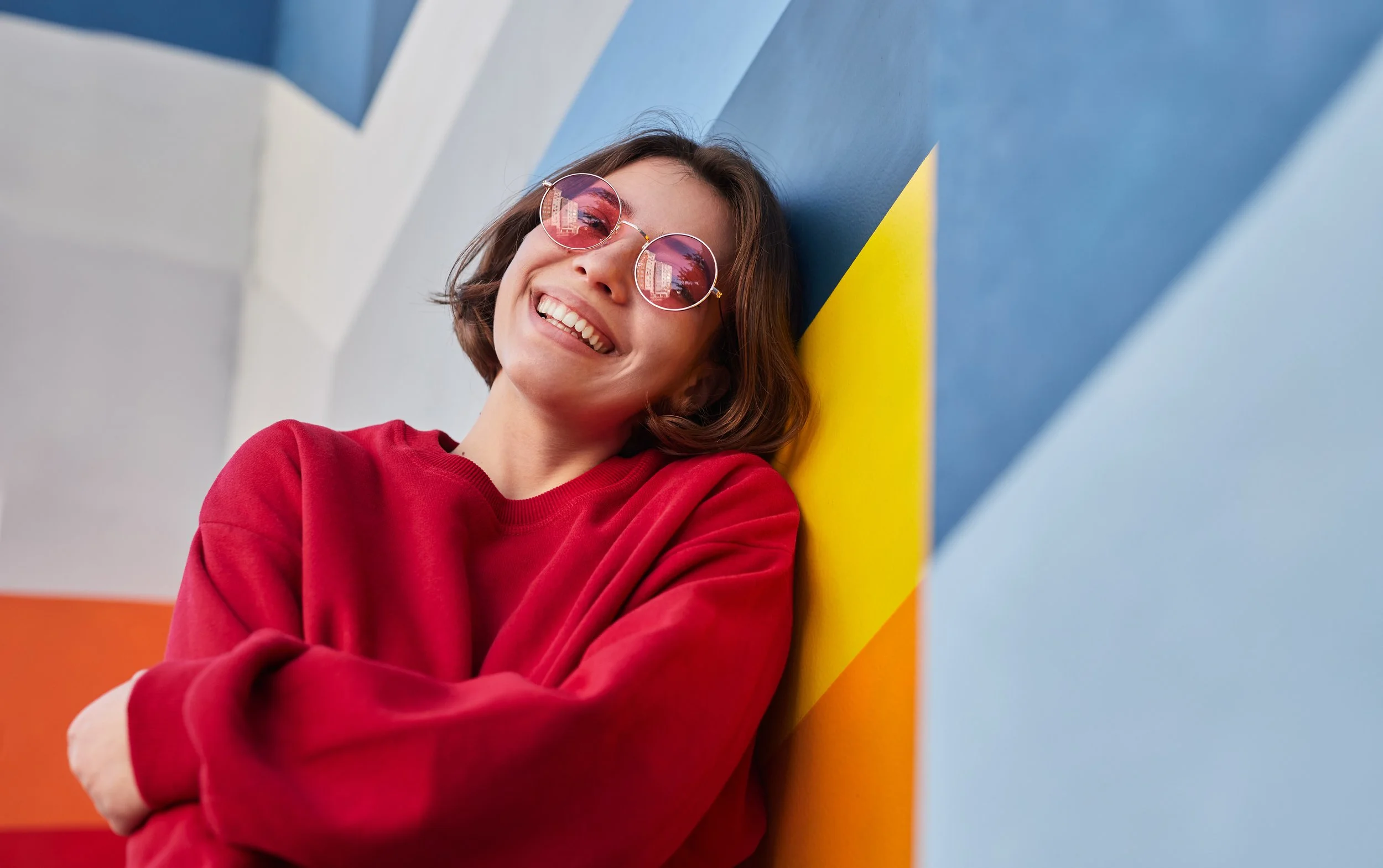 Jeune femme souriante portant des lunettes de soleil roses, vêtue d'un sweat rouge, posant contre un mur coloré composée de sections bleues, jaunes, orange et blanches.