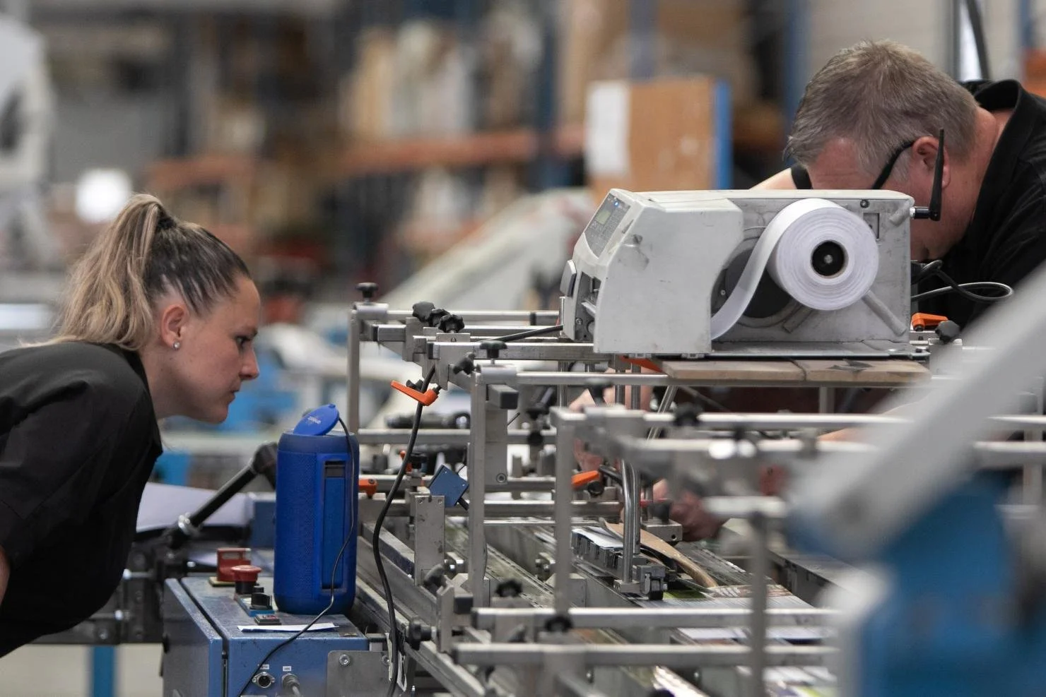 Deux personnes travaillent sur une machine dans un atelier industriel. La femme à gauche regarde attentivement la machine, tandis que l'homme à droite semble concentré sur une tâche spécifique.