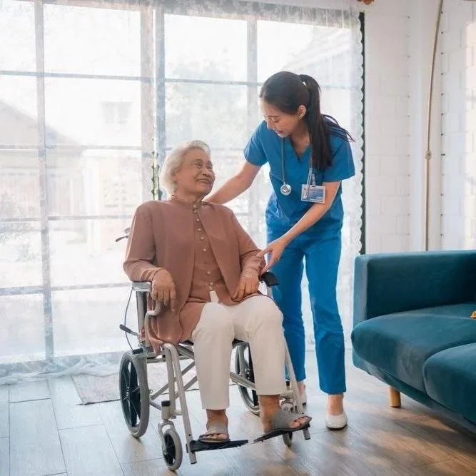 Une infirmière en uniforme bleu souriante aide une femme âgée en fauteuil roulant dans une pièce lumineuse.