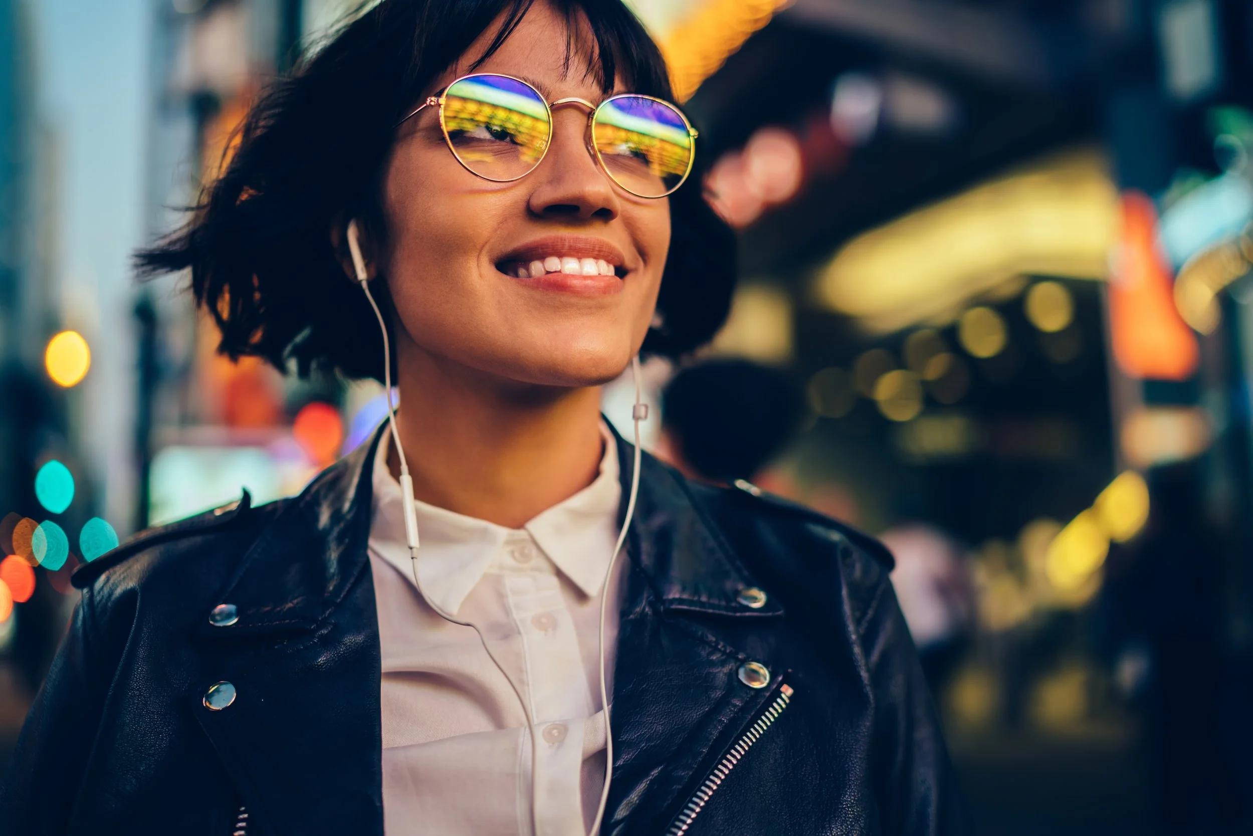 Femme souriante portant des lunettes de soleil colorées, écoutant de la musique avec des écouteurs dans un environnement urbain nocturne illuminé par des lumières colorées.