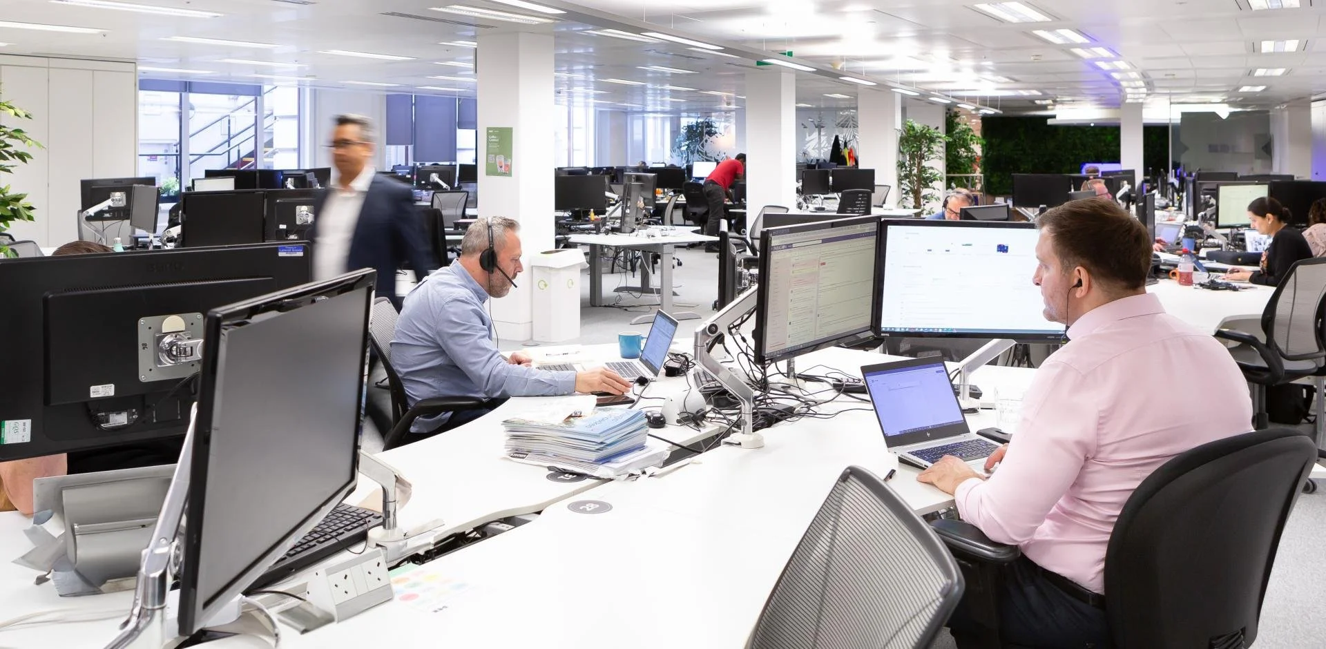 Un espace de bureau ouvert avec plusieurs employés travaillant sur des ordinateurs, certains portés des casques. Des écrans d'ordinateurs et une pile de documents sont visibles sur les bureaux.