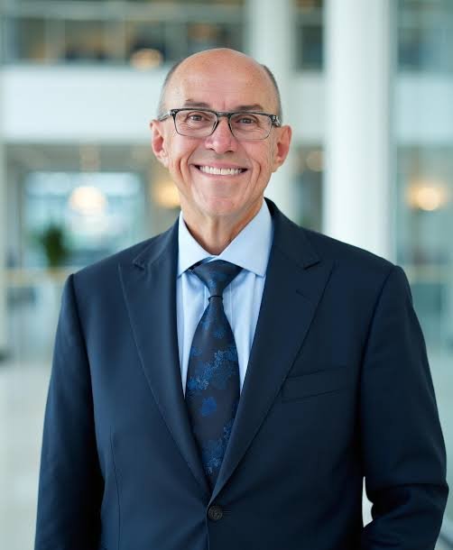 Smiling older man in a dark suit, blue shirt, and patterned tie, standing in a modern office building with glass windows.