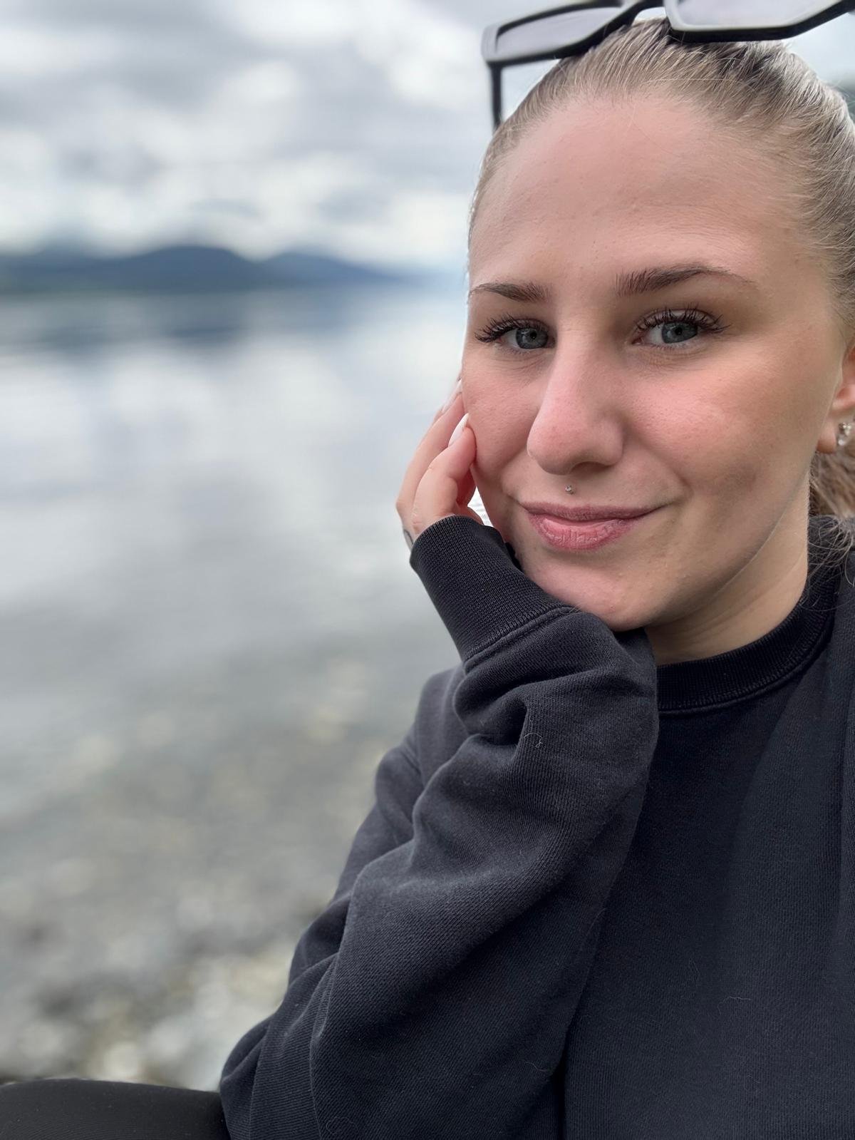 Nahaufnahme eines lächelnden jungen Frauen mit blonden Haaren, blauen Augen, Nasenpiercing, Sonnenbrille auf Kopf, trägt schwarze Kleidung, sitzt an einem see mit Bergen im Hintergrund, bewölkter Himmel.