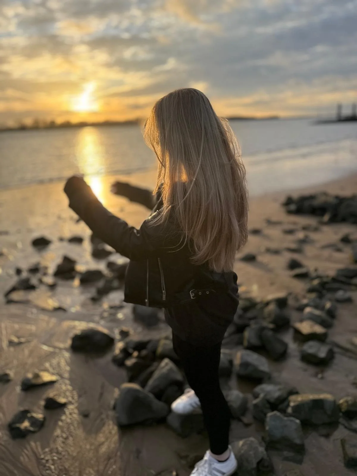 Junge Frau mit langen Haaren, schwarzer Jacke und weißen Turnschuhen steht an einem Strand bei Sonnenuntergang, blickt aufs Wasser, umgeben von Steinen.