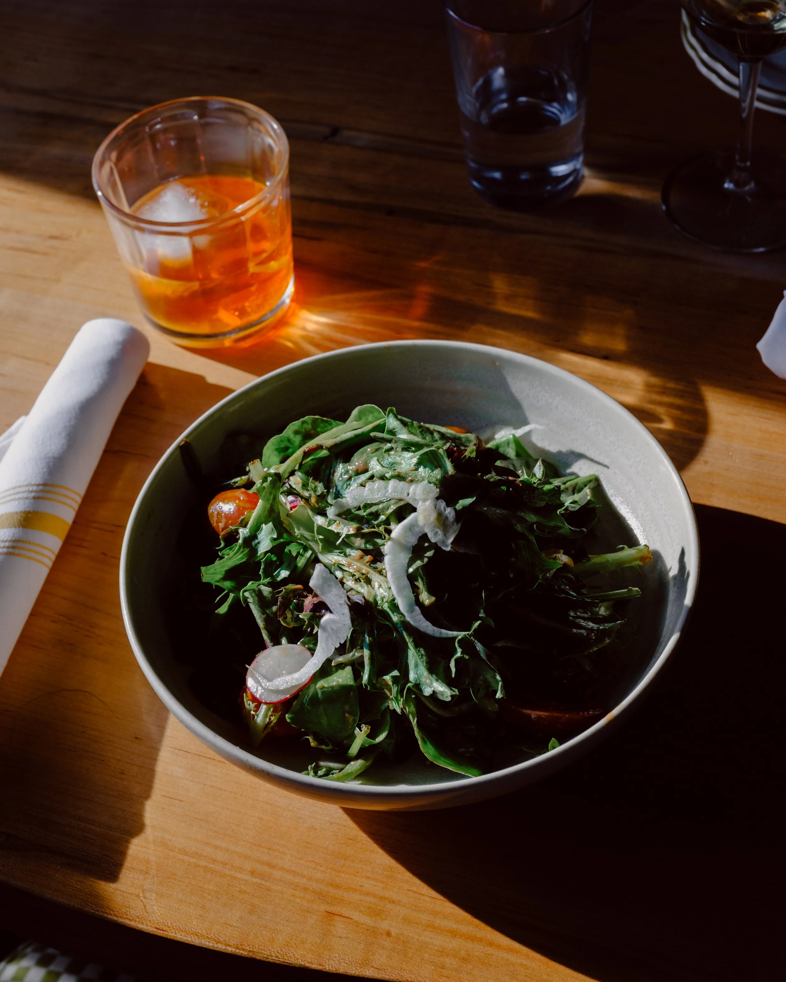 A bowl of fresh mixed greens salad with cherry tomatoes and shaved cheese on a wooden table, illuminated by natural light from an unseen window. In the background, there are three drinking glasses, one filled with an orange beverage and ice, and two 