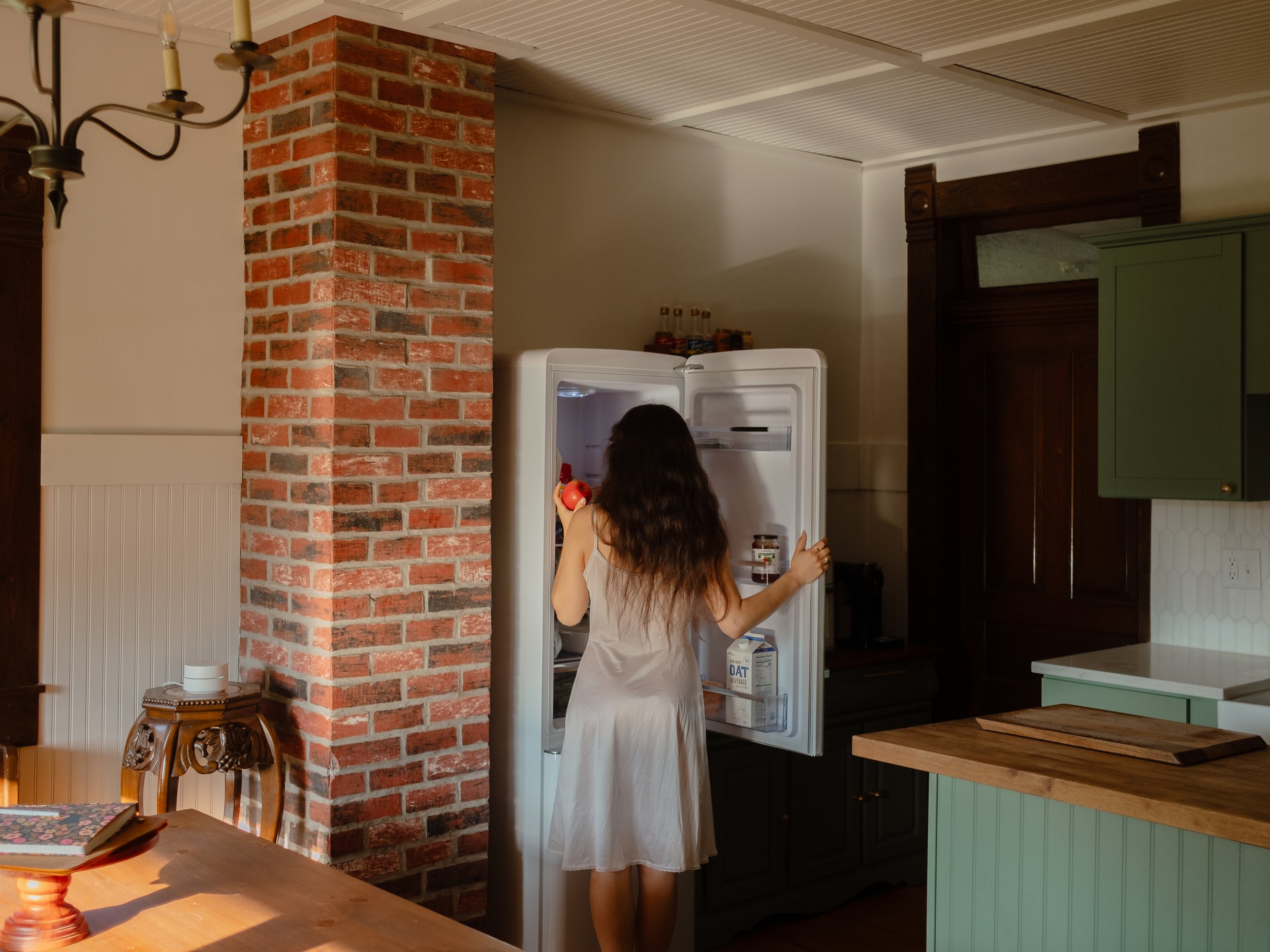 A woman with long dark hair wearing a beige dress is standing in front of an open refrigerator, reaching inside to get an apple. The kitchen has a brick pillar, a wooden table with a decorative candle stand, and green and dark wooden cabinetry.