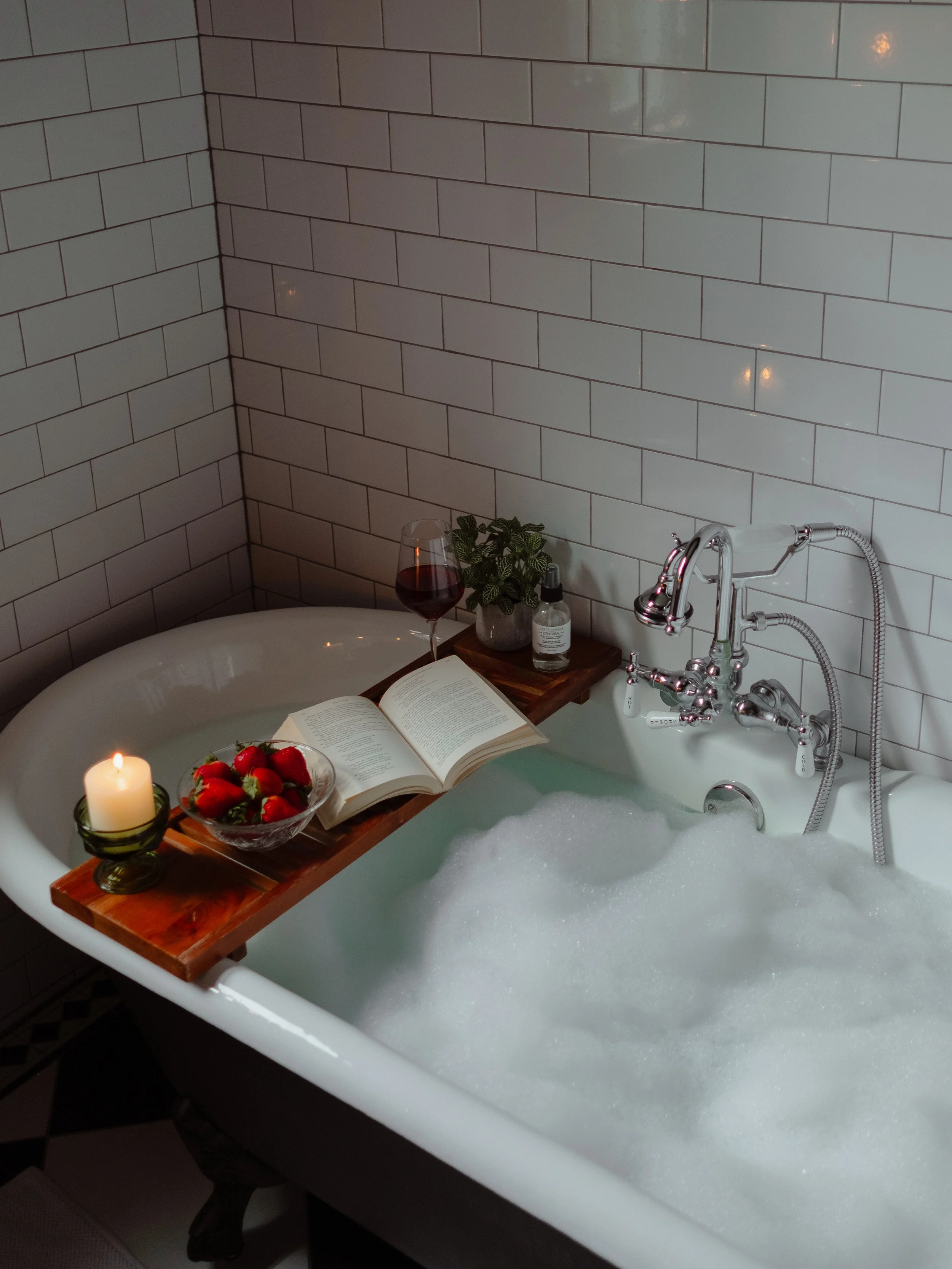 A bathtub filled with bubbles, with a candle and a bowl of strawberries on a wooden tray on the edge. There is a glass of red wine, a potted plant, a book, and a bottle of lotion nearby. The background features white subway tile walls and a vintage-s