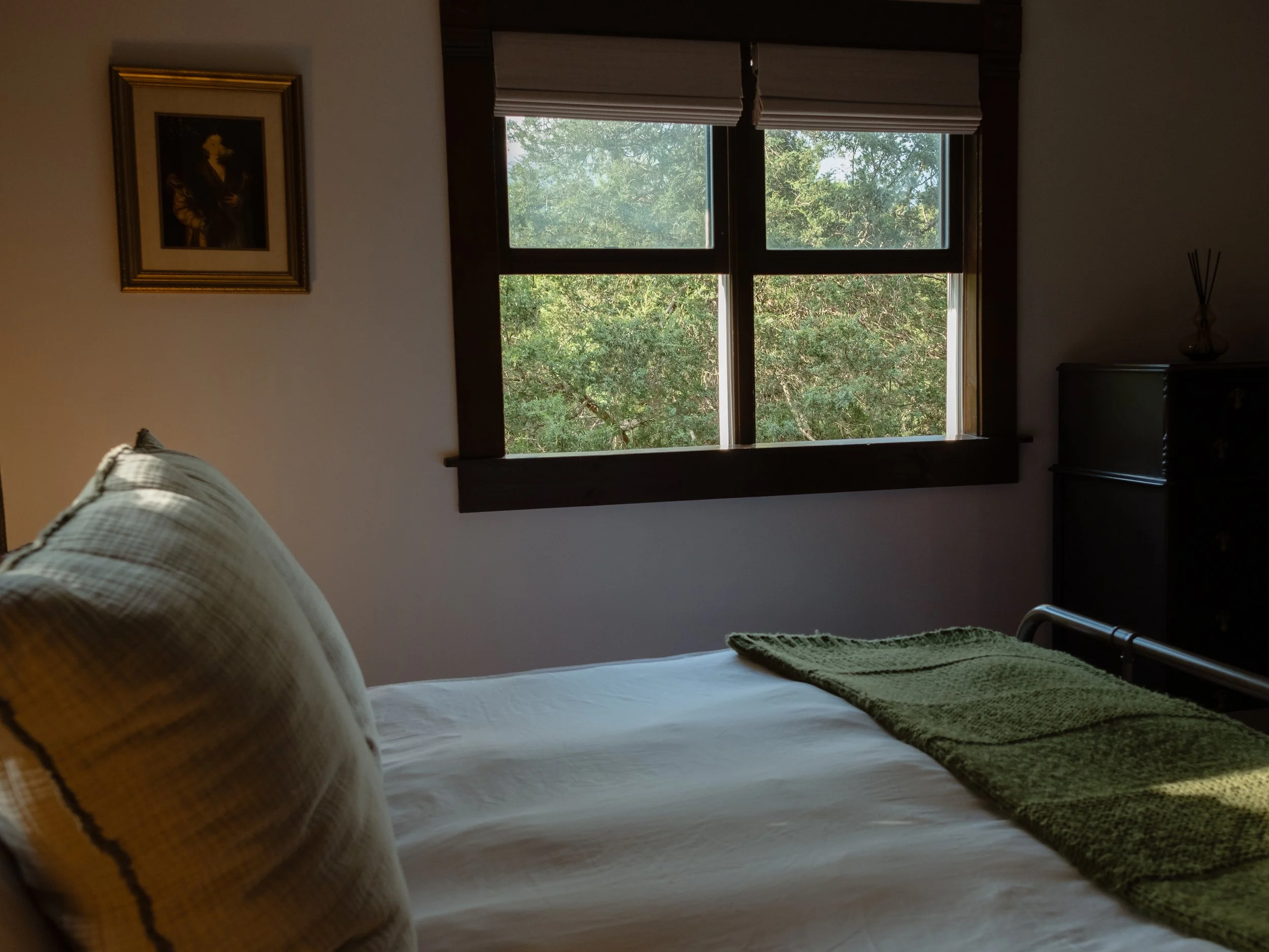 A bedroom with a bed, a window showing green trees outside, and a framed portrait on the wall.