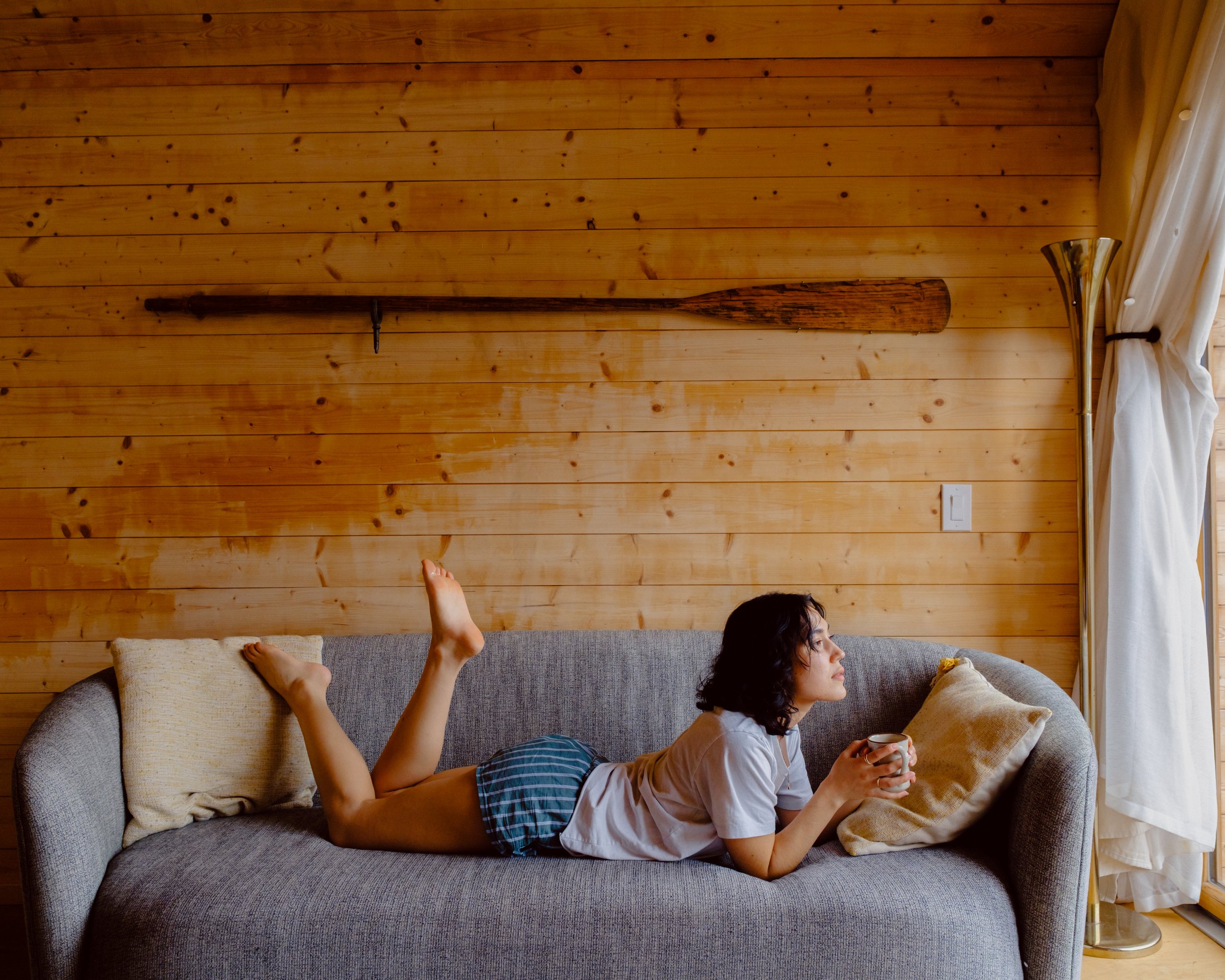 A woman lying on a gray couch, holding a coffee mug, with a child’s foot and hand visible, in front of a wooden wall with a curtain and floor lamp.