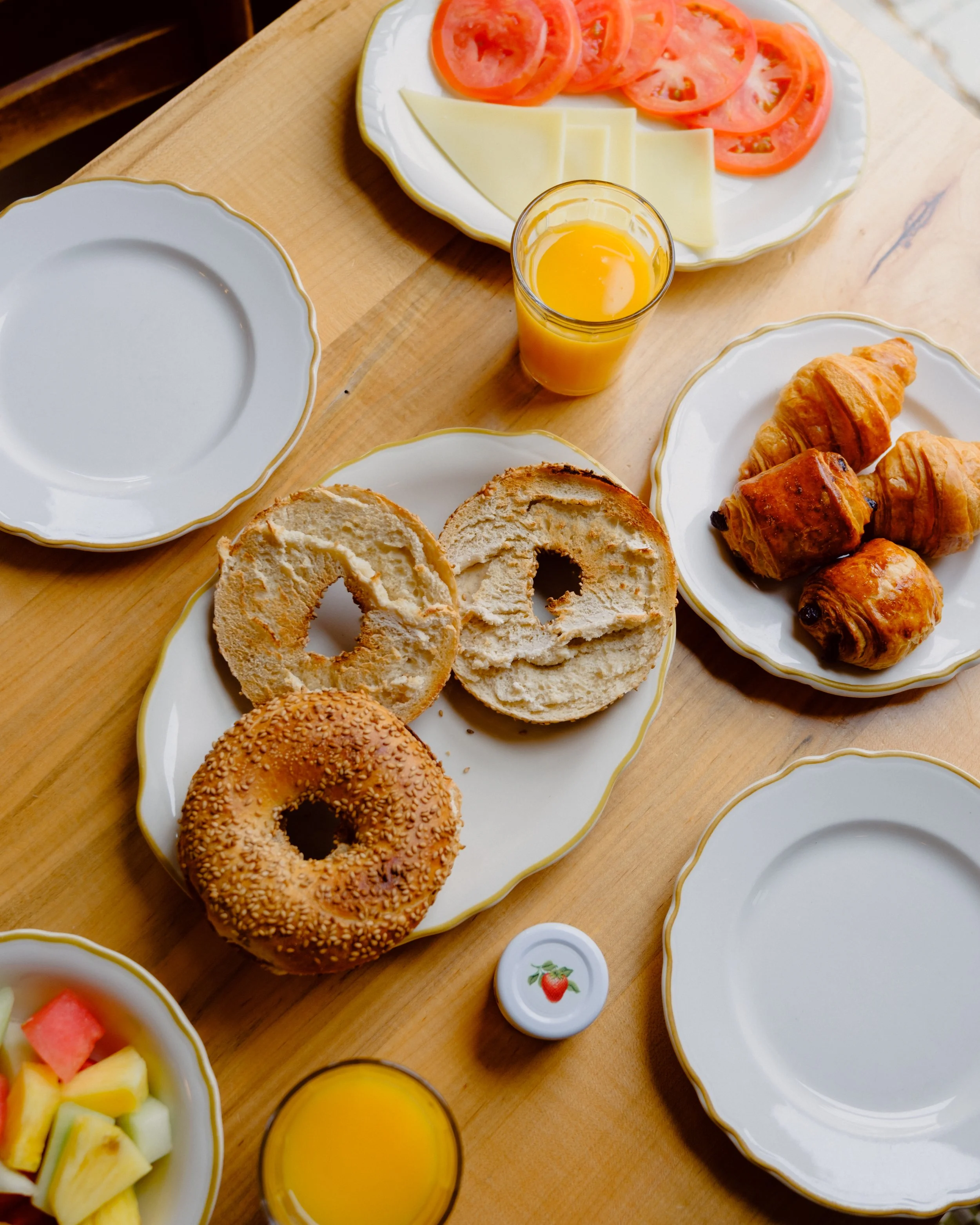 Breakfast spread on a wooden table including sliced tomatoes and cheese on a plate, a glass of orange juice, a bagel with sesame seeds, mixed fruit salad, croissants, and empty plates.