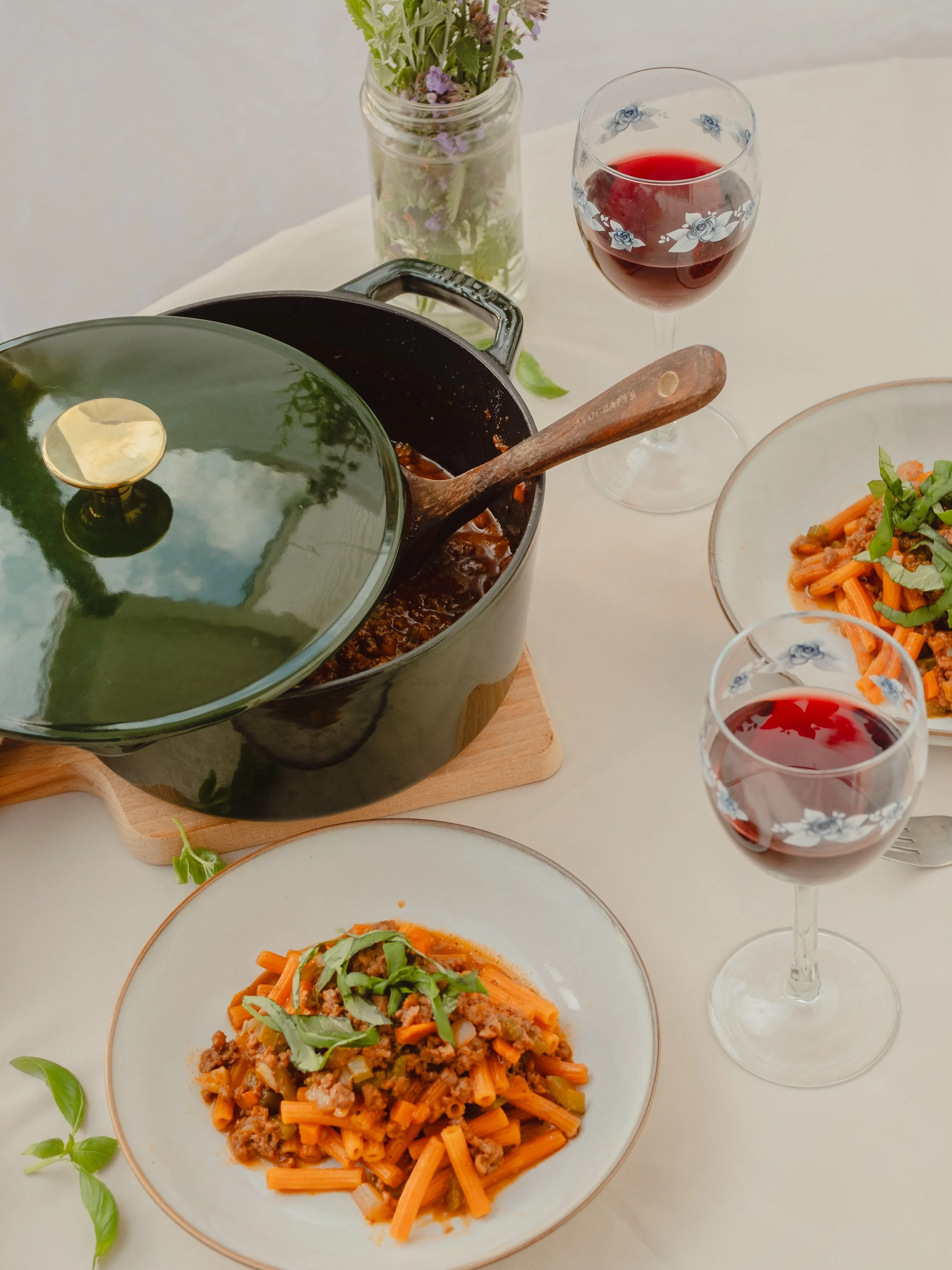 A plate of spaghetti with tomato sauce and greens, two glasses of red wine, a pot with a lid, a glass jar with flowers, and a bowl with a pasta dish on a white table.
