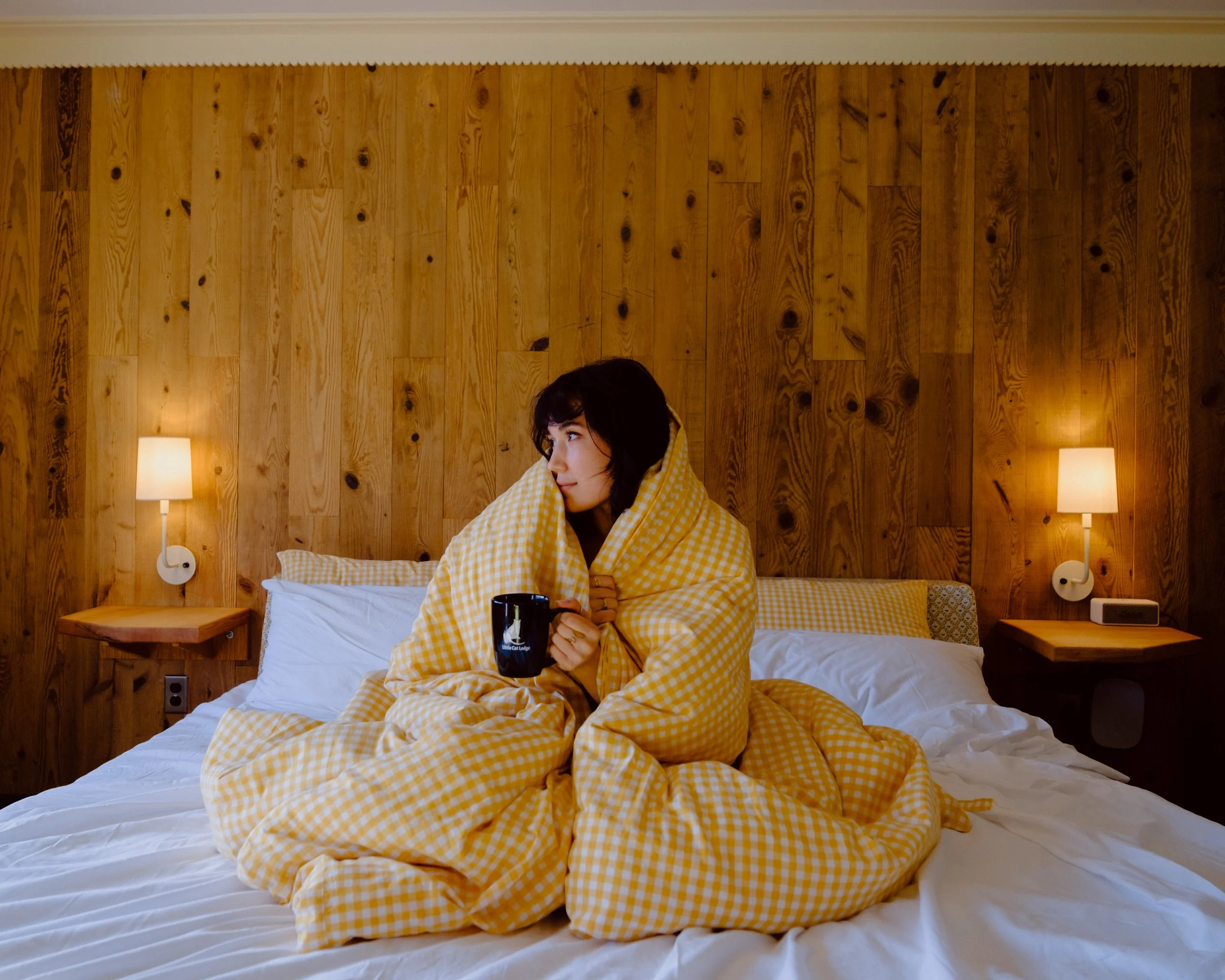 A woman sitting on a bed with yellow checkered blanket, holding a black mug, in a room with wooden paneled wall and two wall-mounted lamps.