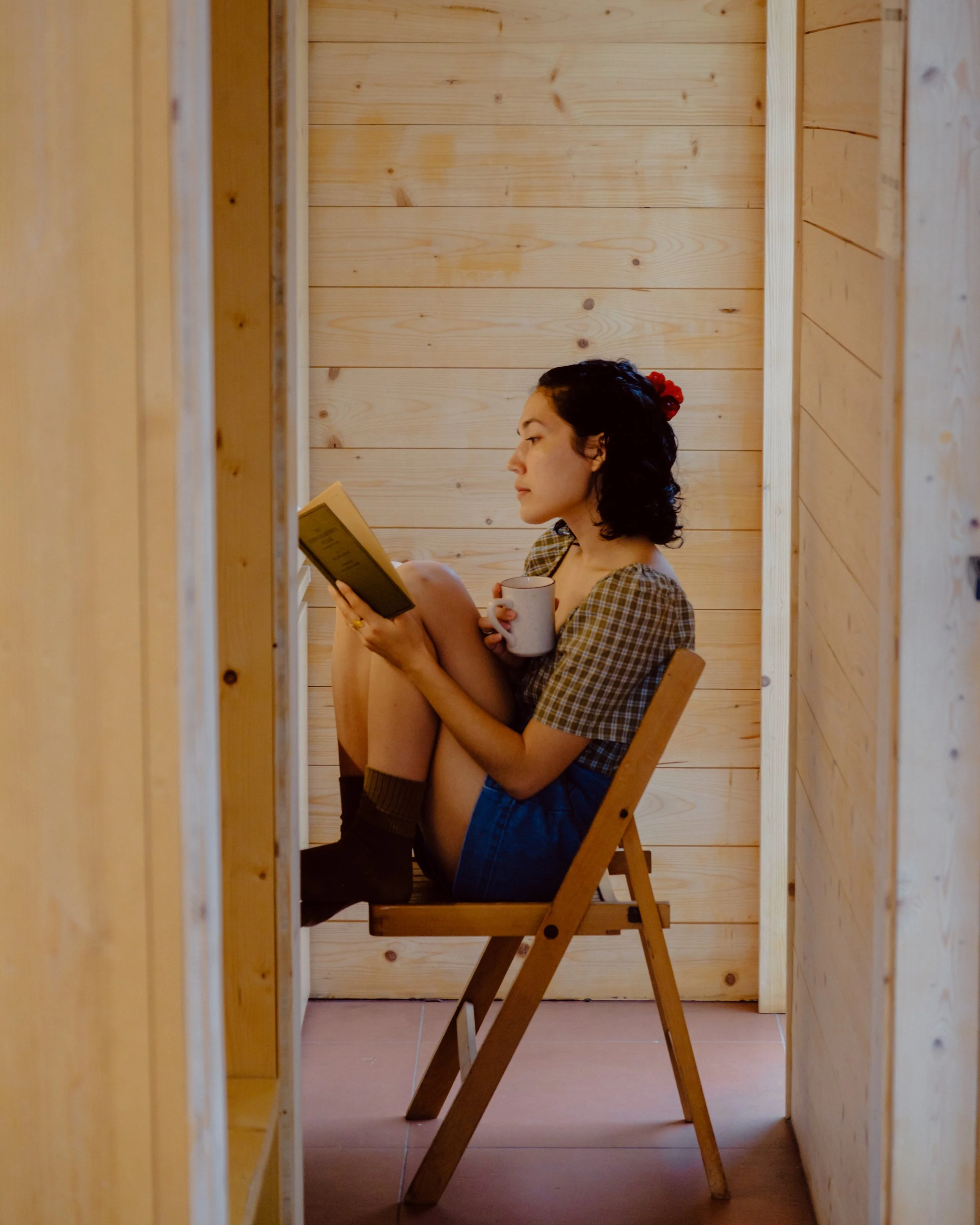 A woman with curly dark hair, wearing a plaid shirt, sitting on a wooden chair in a cozy, wood-paneled room, reading a book and holding a cup.