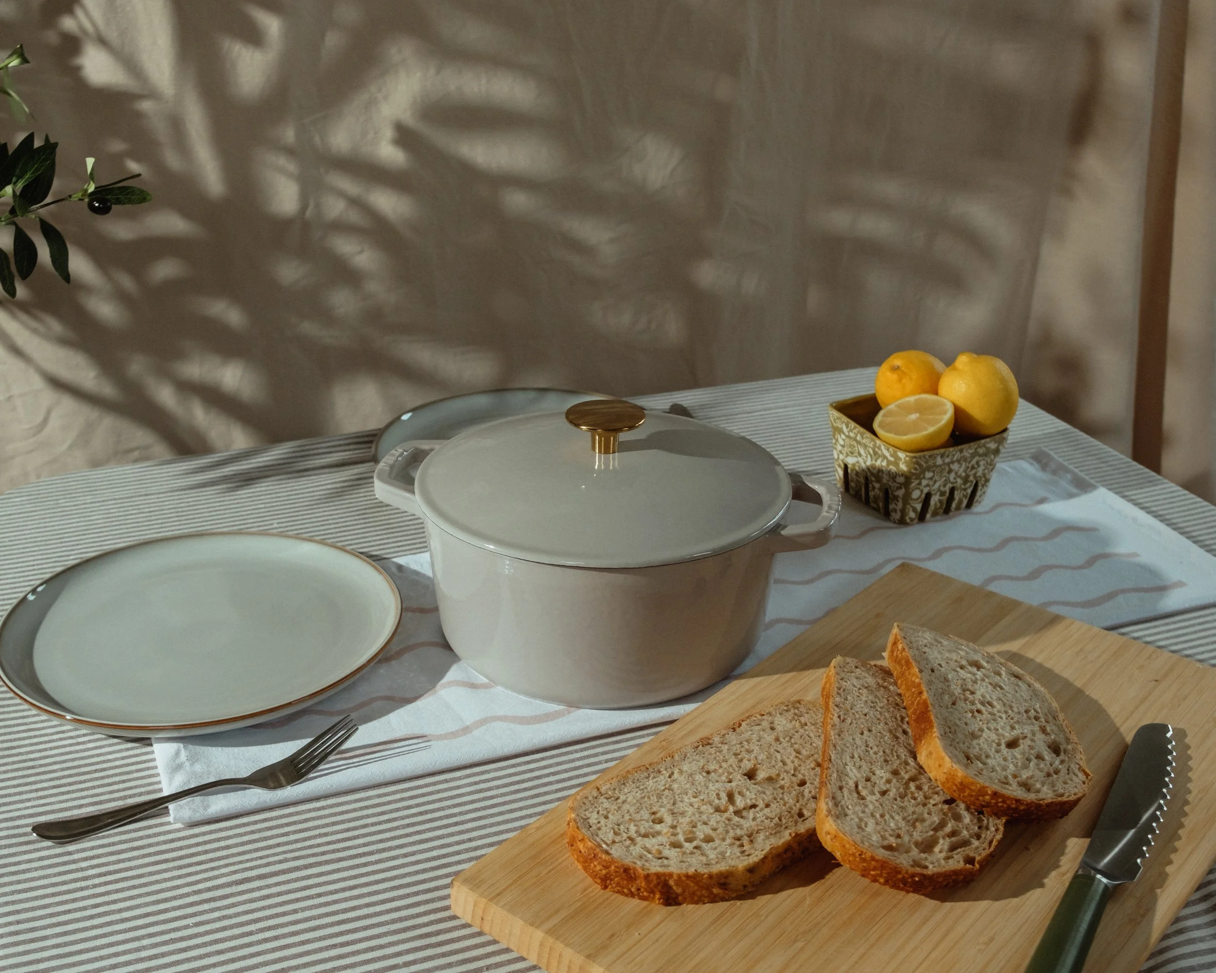 A table with slices of bread on a wooden cutting board, a white covered dish, a plate, a fork, and a knife. In the background, a bowl with whole and halved lemons and a decorative bowl. Green leaves with black berries are on the left side, casting sh