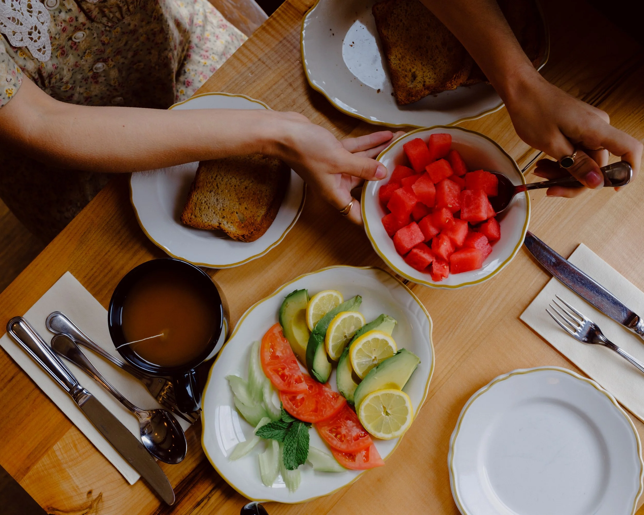 A wooden table with plates of chopped watermelon, sliced cucumbers, tomatoes, lemons, and lettuce, along with some toasted bread, a lemon wedges, and utensils