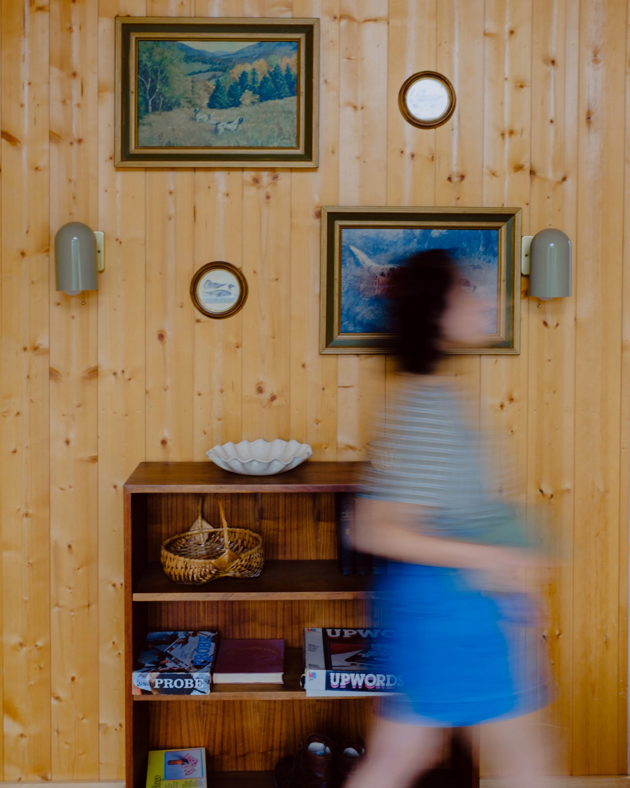 A wooden bookshelf with three shelves, containing board games, a basket, framed artwork, and decorative items. The top shelf has framed landscape paintings and a small circular mirror. The middle shelf holds books, board games labeled 'PROBE' and 'UP