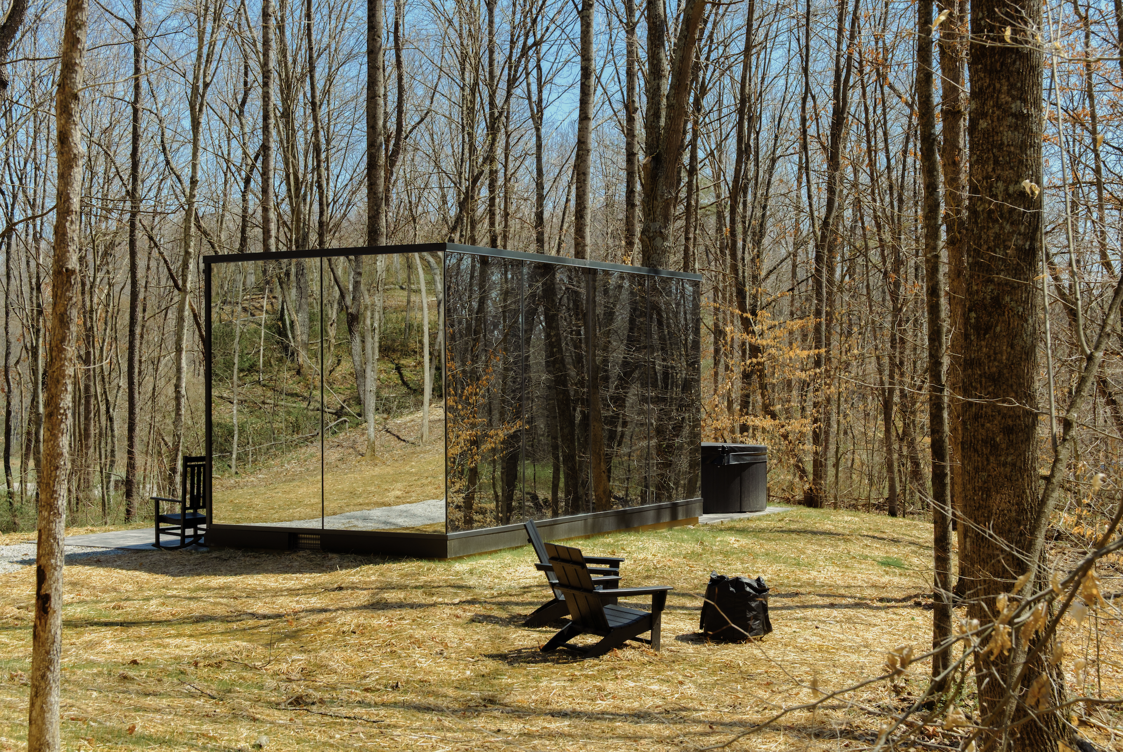 A small outdoor shelter with reflective walls in a wooded area during daytime, with chairs and a trash can nearby.