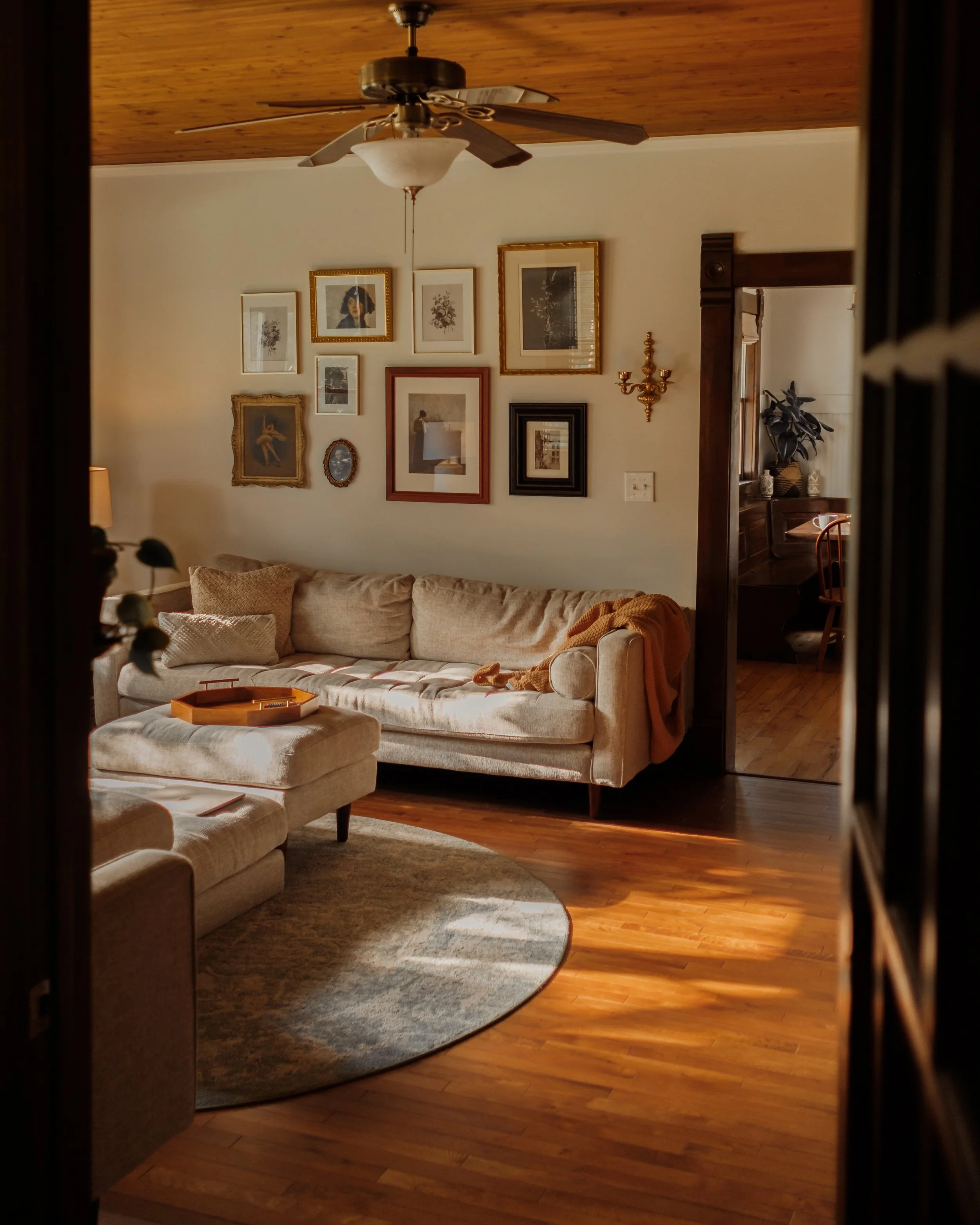 A cozy living room with a beige sofa with pillows, a small ottoman, a wall filled with framed pictures and artwork, a ceiling fan, a doorway leading to another room with a table and chair, and hardwood floors with a circular rug.
