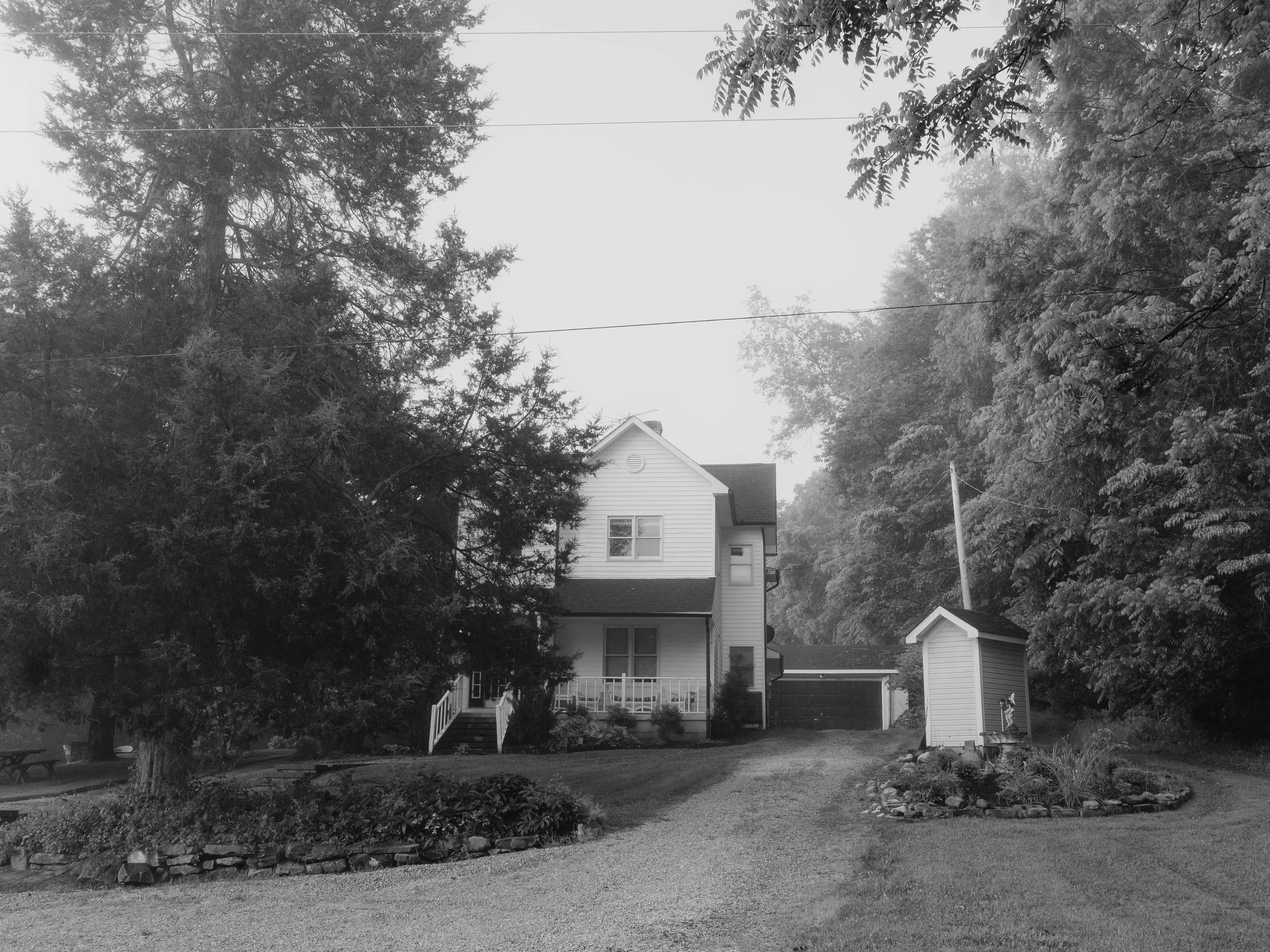 A black and white photo of a suburban house with multiple two and a half stories, including a porch, surrounded by mature trees, a lawn, and a paved pathway leading to the house.