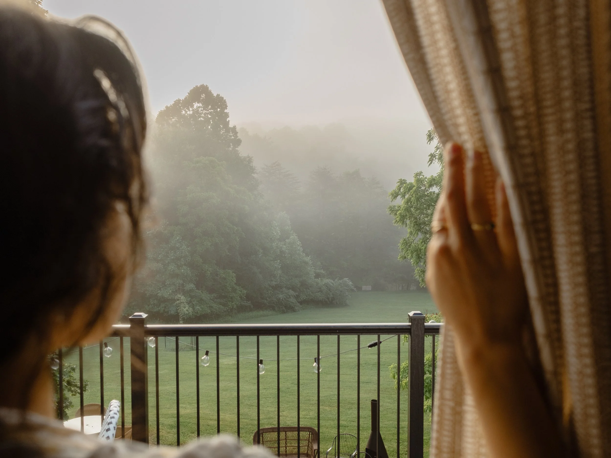 View of a foggy landscape with trees, seen through a window framed by curtains, possibly from a balcony.