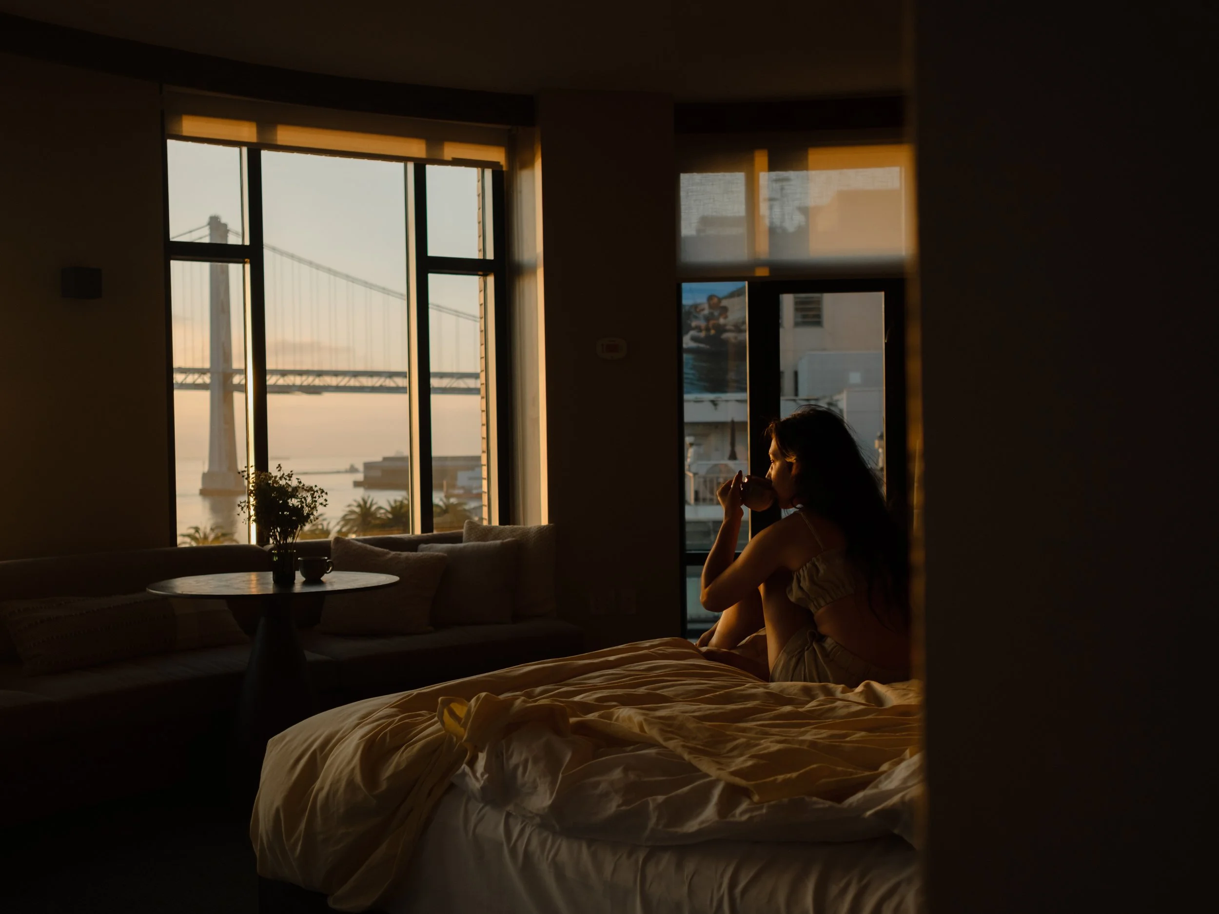 A woman sitting on a bed, drinking from a cup, in a dimly lit room with large windows showing a bridge and cityscape outside during sunset.