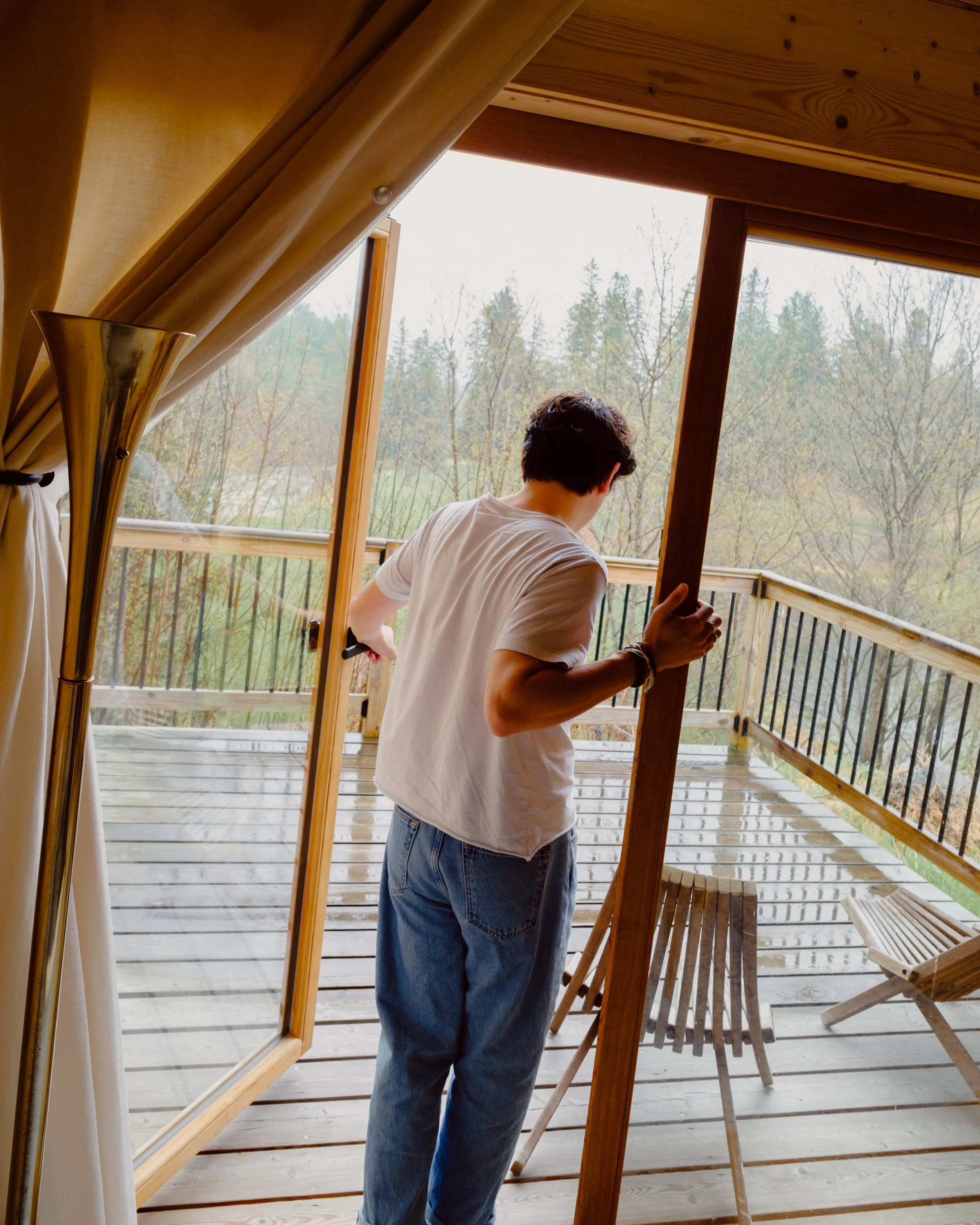 A person with short dark hair, wearing a white t-shirt and blue jeans, is standing on a wooden deck, working on a wooden structure near the railing, with a scenic view of trees and foggy weather outside.