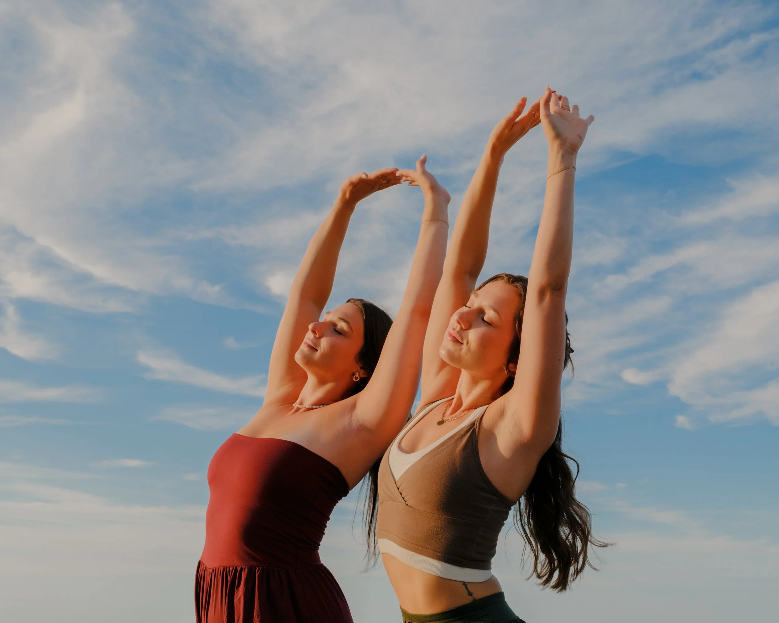 Three women practicing yoga outdoors with arms raised in front of a blue sky with clouds.