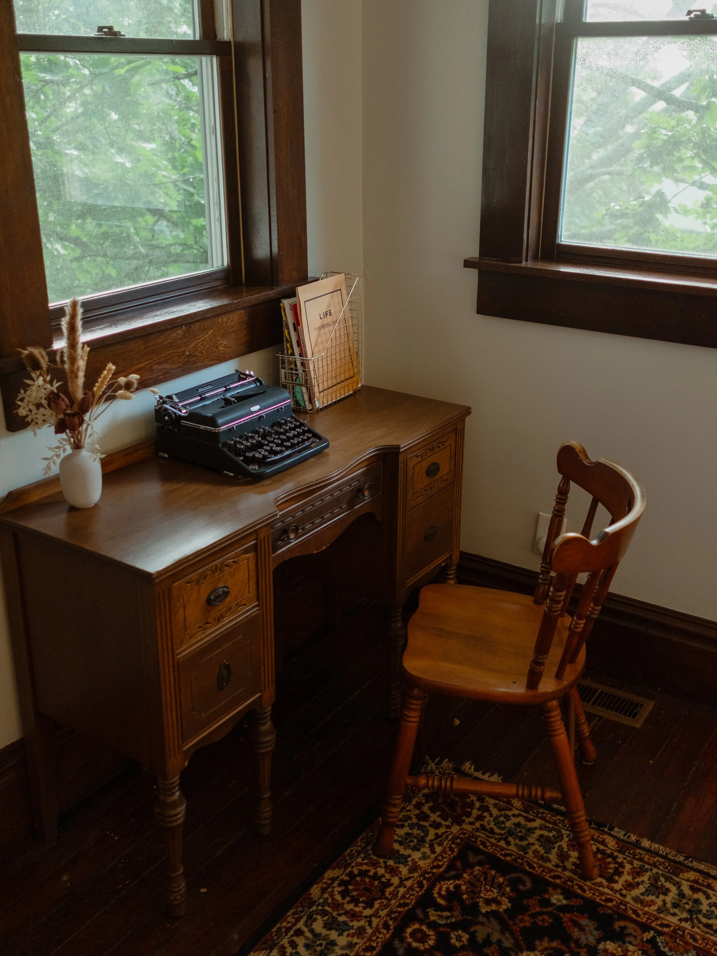 Interior of a room with a wooden desk near two windows with dark wood trim. On the desk, there is a black typewriter, a wire bookshelf with books and a file folder labeled 'LIFE'. A wooden chair with turned spindles is in front of the desk, and a pat