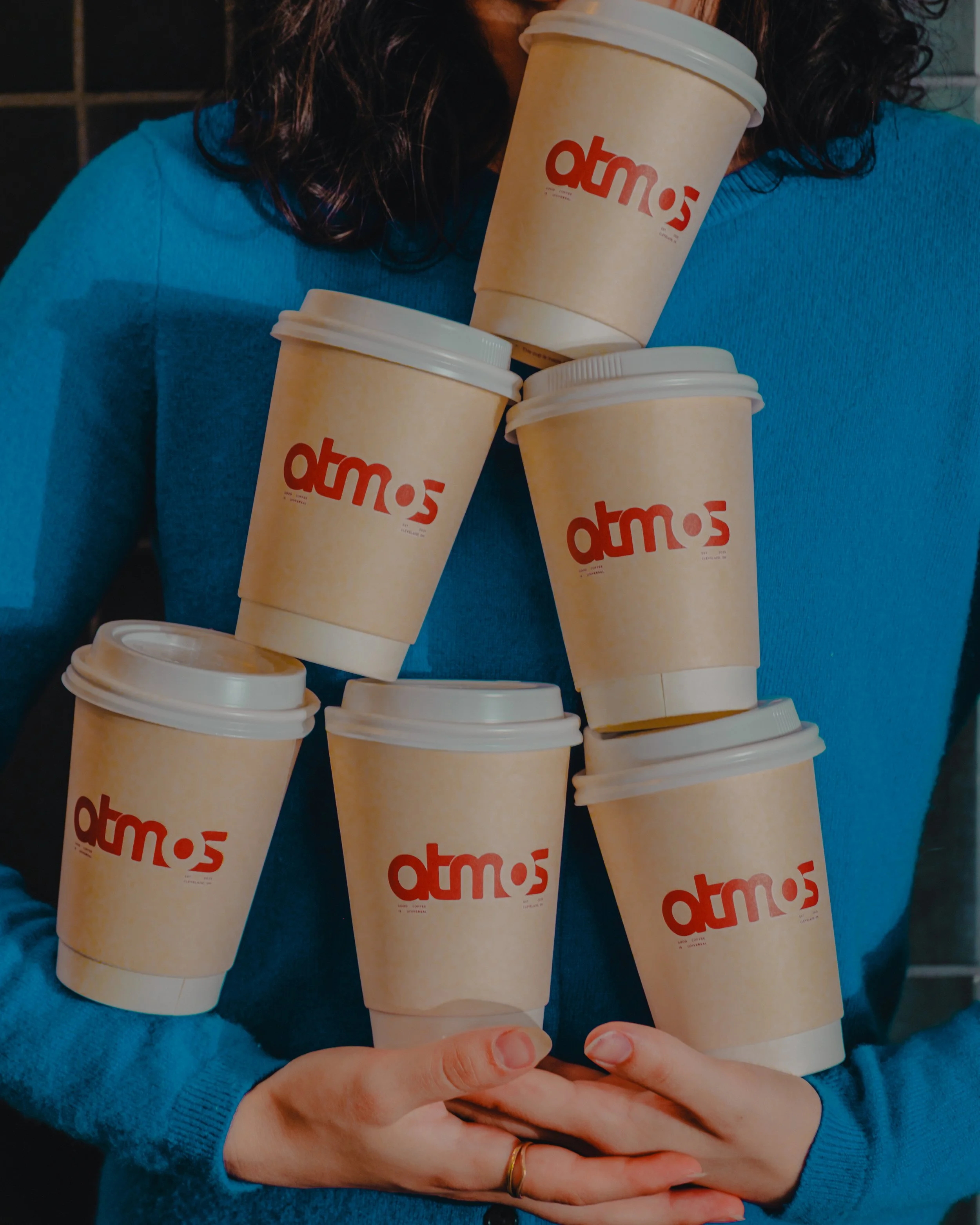 Person wearing a blue sweater holds multiple cold beverage cups from atmós, forming a stack.