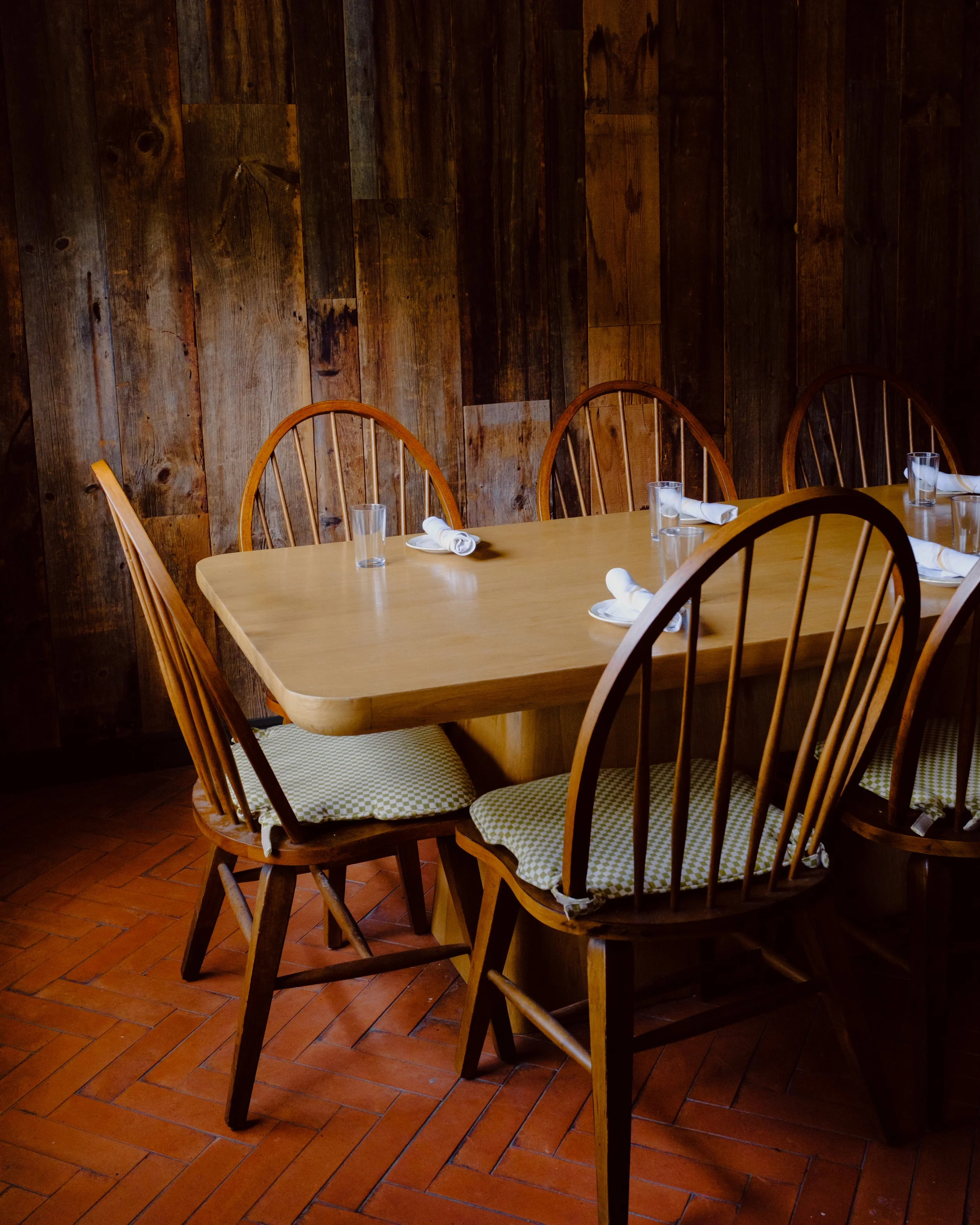 A wooden dining table with chairs in a rustic indoor setting. The table has white cloth napkins and drinking glasses, some upright and some on plates. The background features a dark brown wooden wall and a terracotta tiled floor.