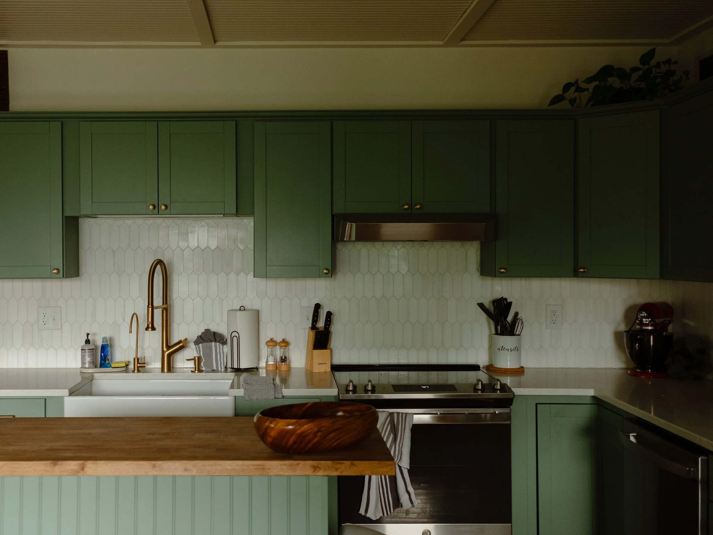 Green kitchen cabinets above a white countertop with a sink and gold faucet. Various kitchen utensils and items are on the counter, including soap bottles, a towel, and a wooden bowl on a wooden island in the foreground. The wall behind has a white t