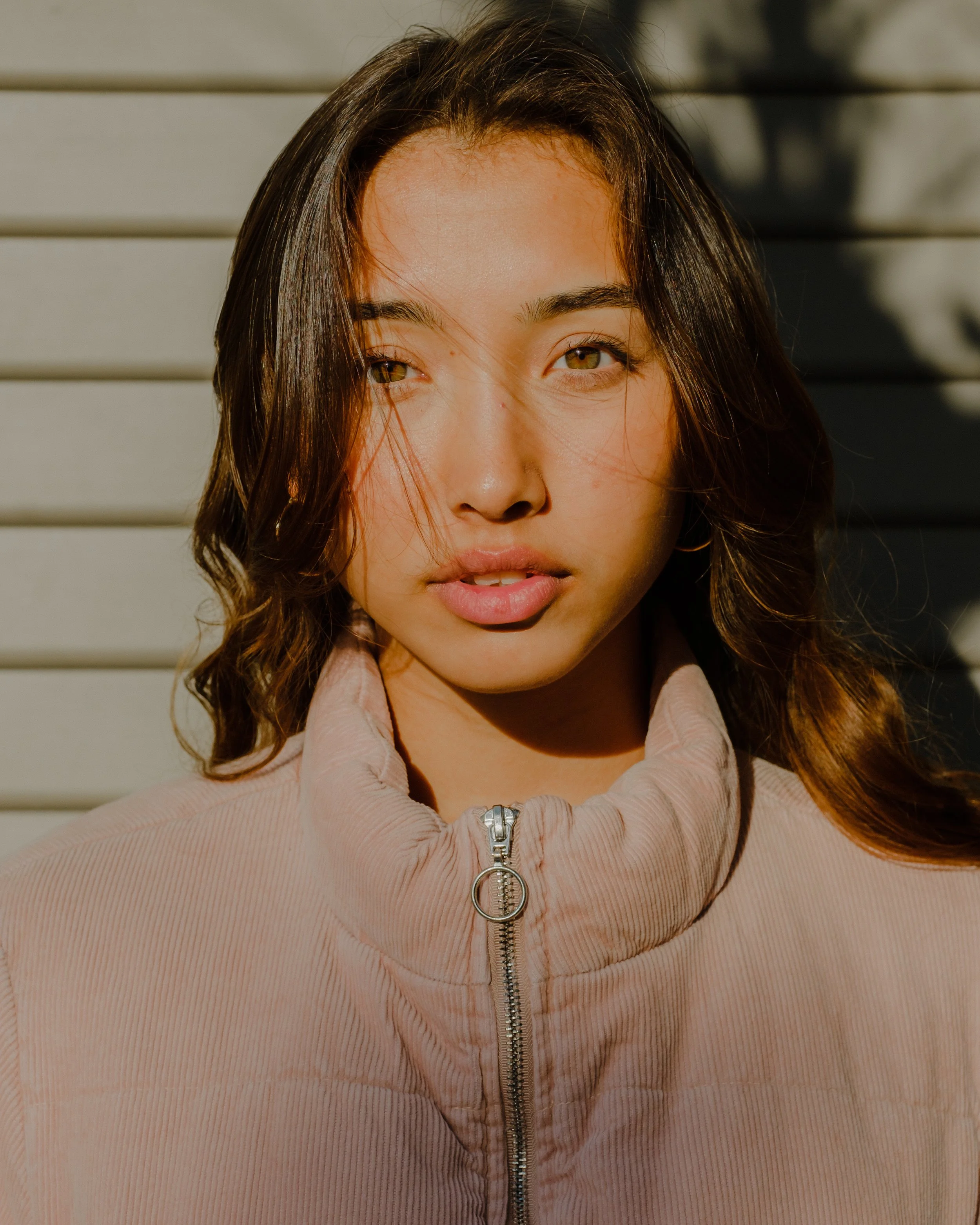 A woman with brown hair and hazel eyes is wearing a beige zip-up jacket with a ring pull zipper and a collared shirt underneath. She is outdoors, standing in front of a gray horizontal slat wall, with sunlight highlighting her face and hair.