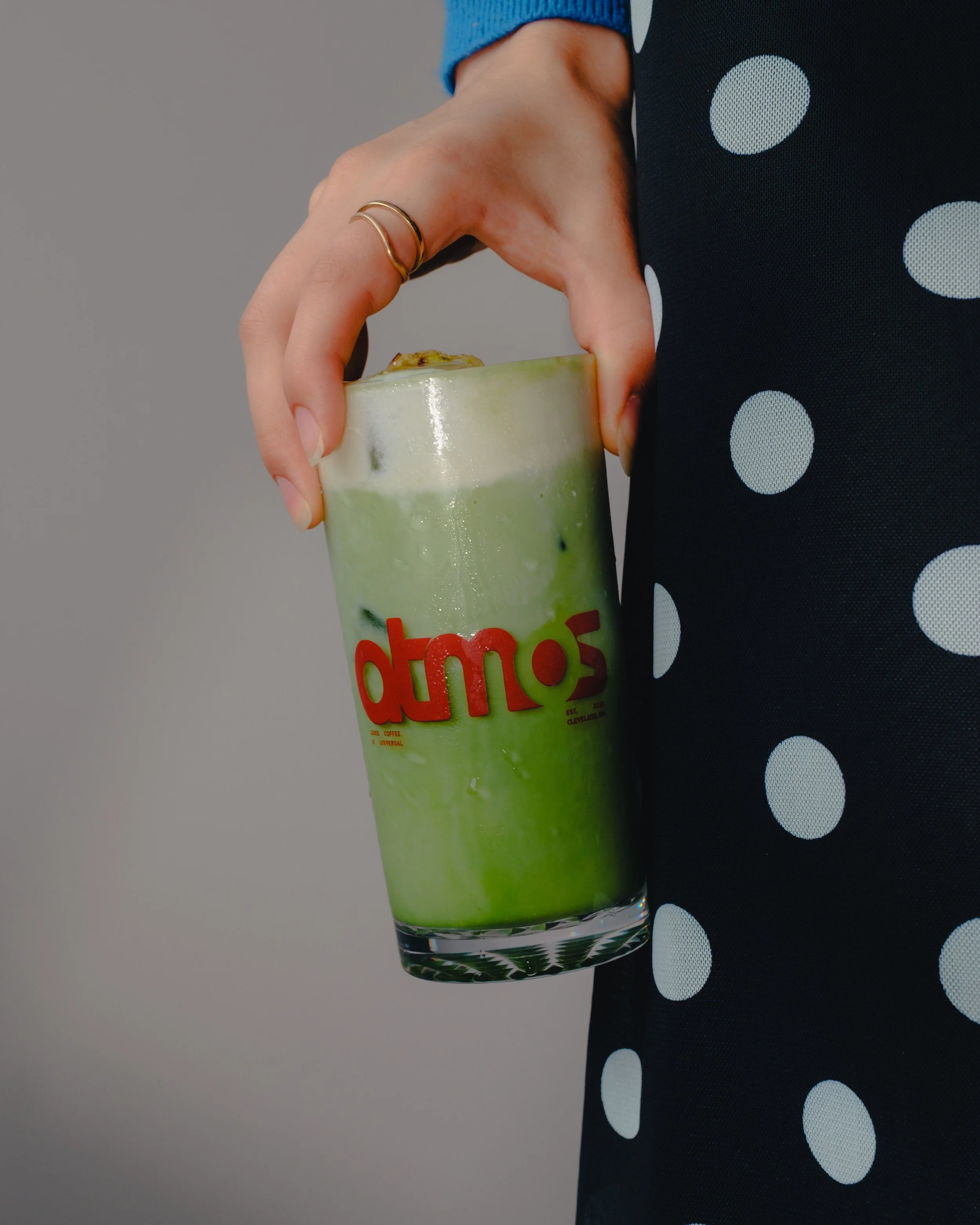 A person wearing a blue sleeve and black polka-dot dress is holding a glass of green beverage with ice and a garnish on top.