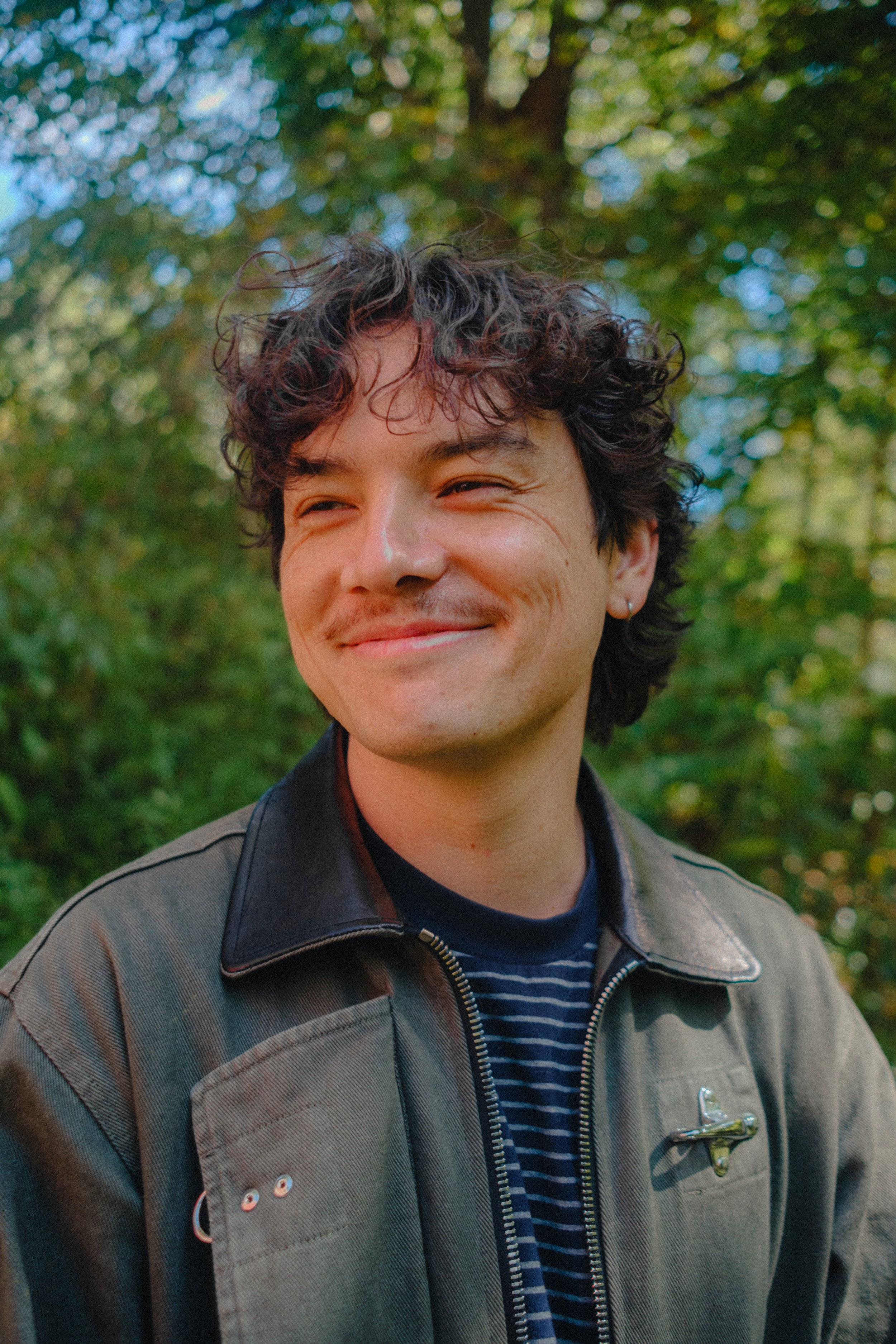 A young man with curly dark hair, smiling, wearing a gray jacket with black leather accents and a striped shirt, standing outdoors with blurred green trees in the background.