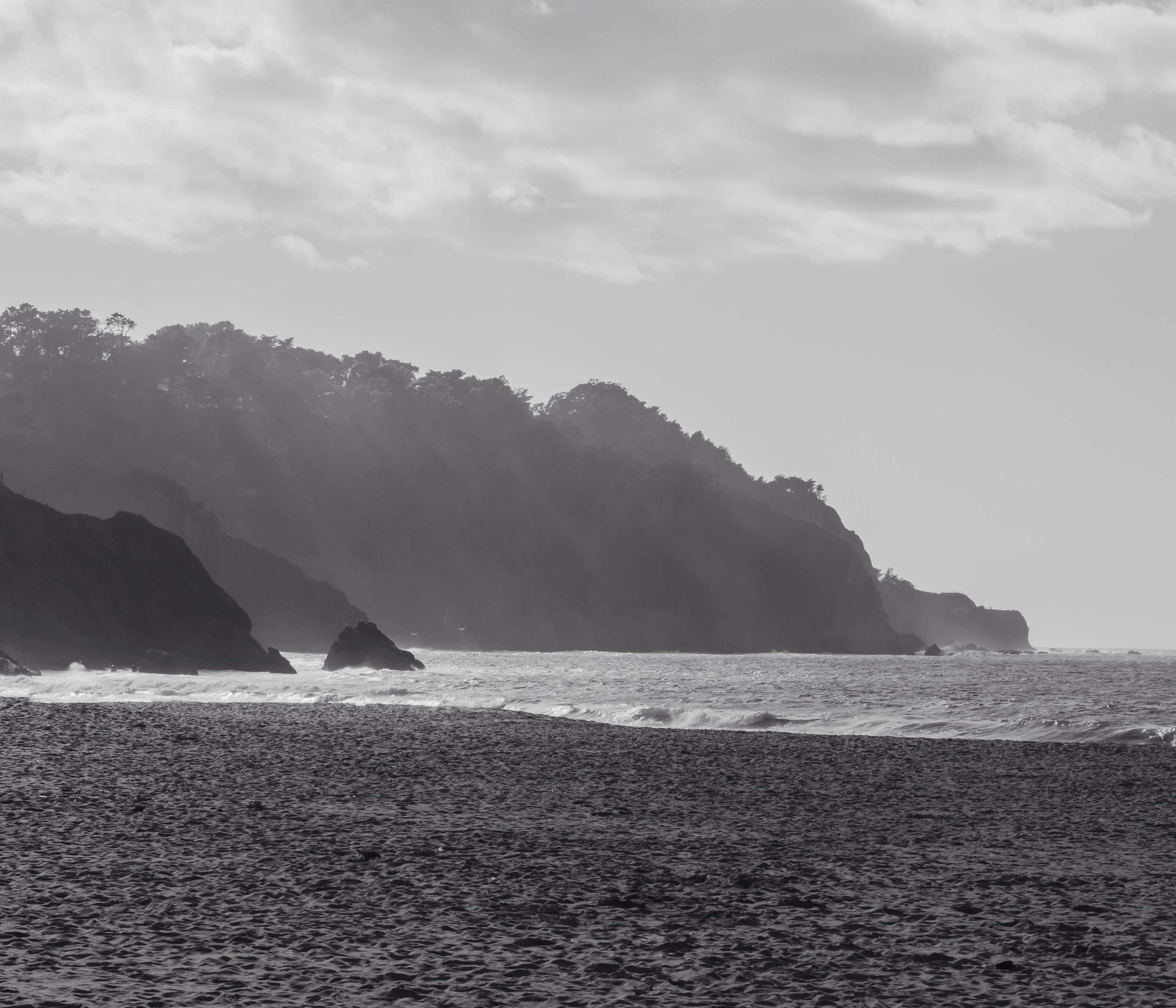 Black and white photograph of a rocky coastline with trees on the hills and turbulent ocean waves crashing against the rocks during cloudy weather.