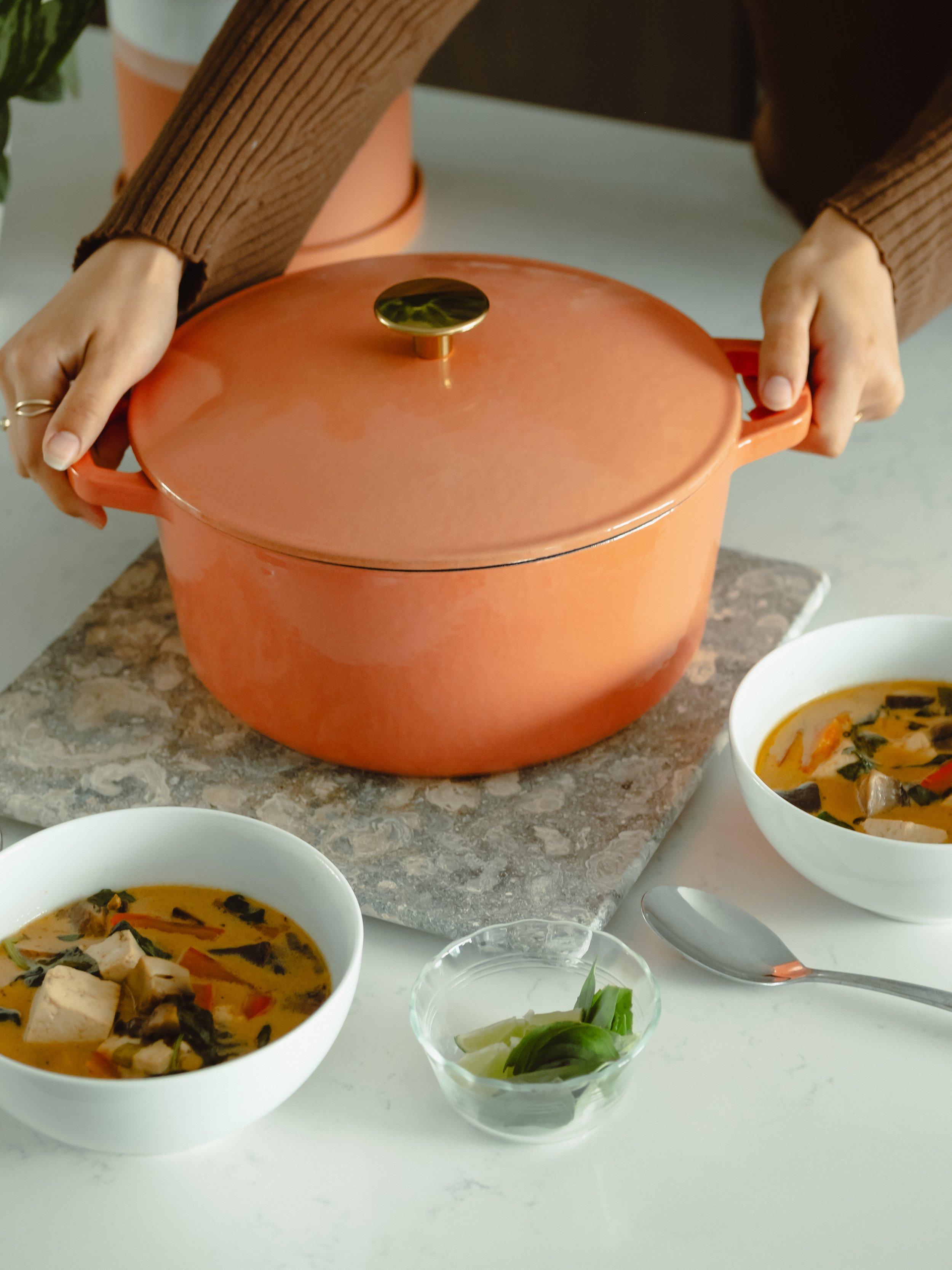Person lifting a large orange pot covered with a lid, with bowls of soup and condiments on a white table.