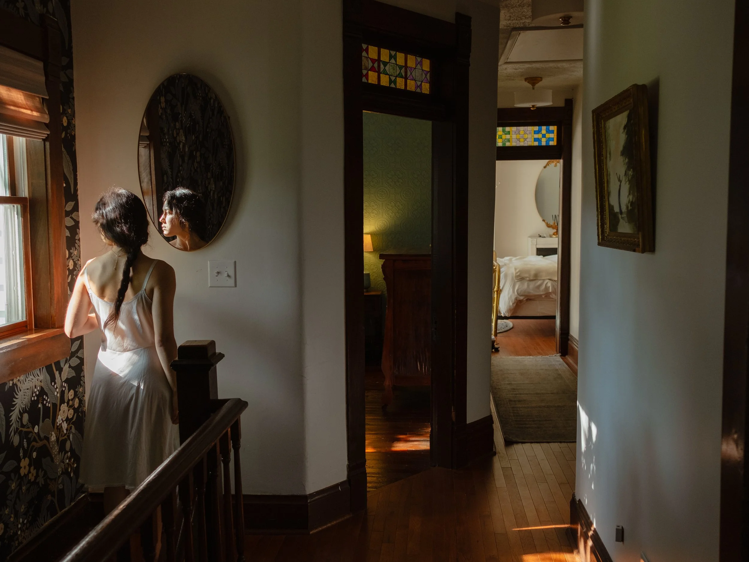 A woman with long dark hair in a braid, wearing a white dress, standing by a window with sunlight streaming in, looking at her reflection in a round mirror on the wall.
