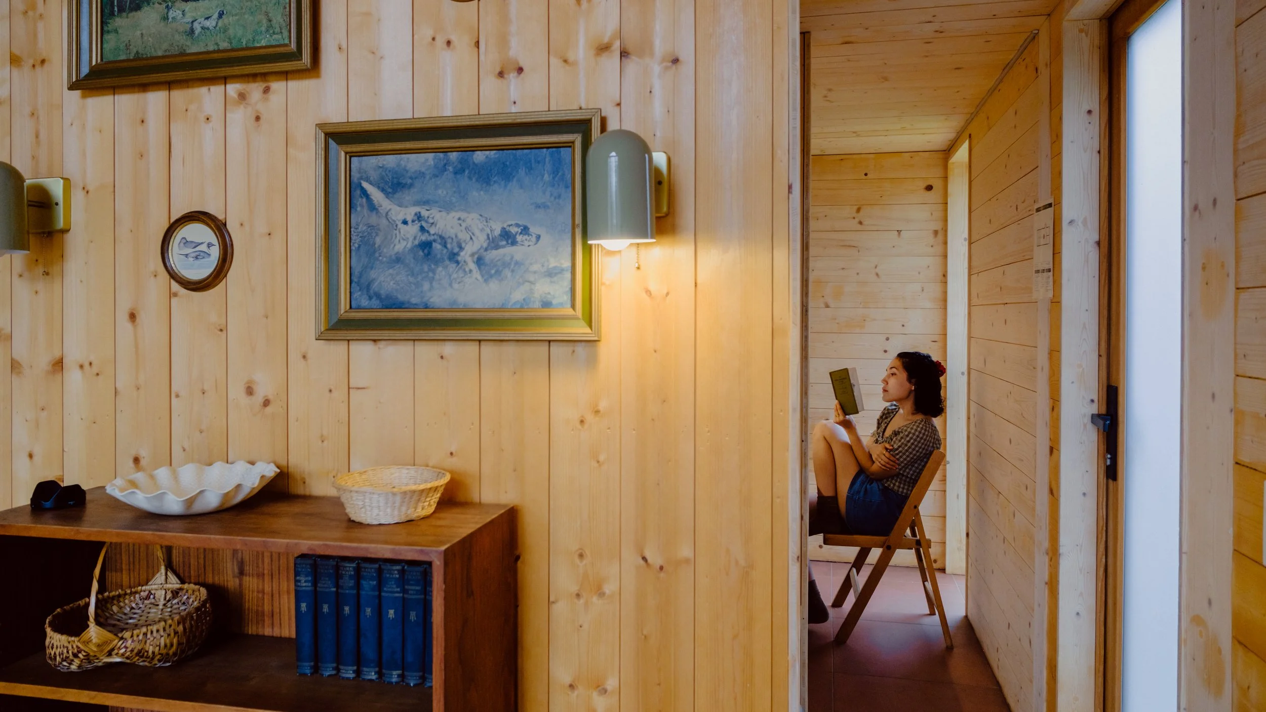 A young woman sitting on a wooden chair in a room with wooden paneled walls, reading a book. There are framed pictures of mountain scenes and a fish on the walls, and a table with a white decorative bowl, a basket, and some books.