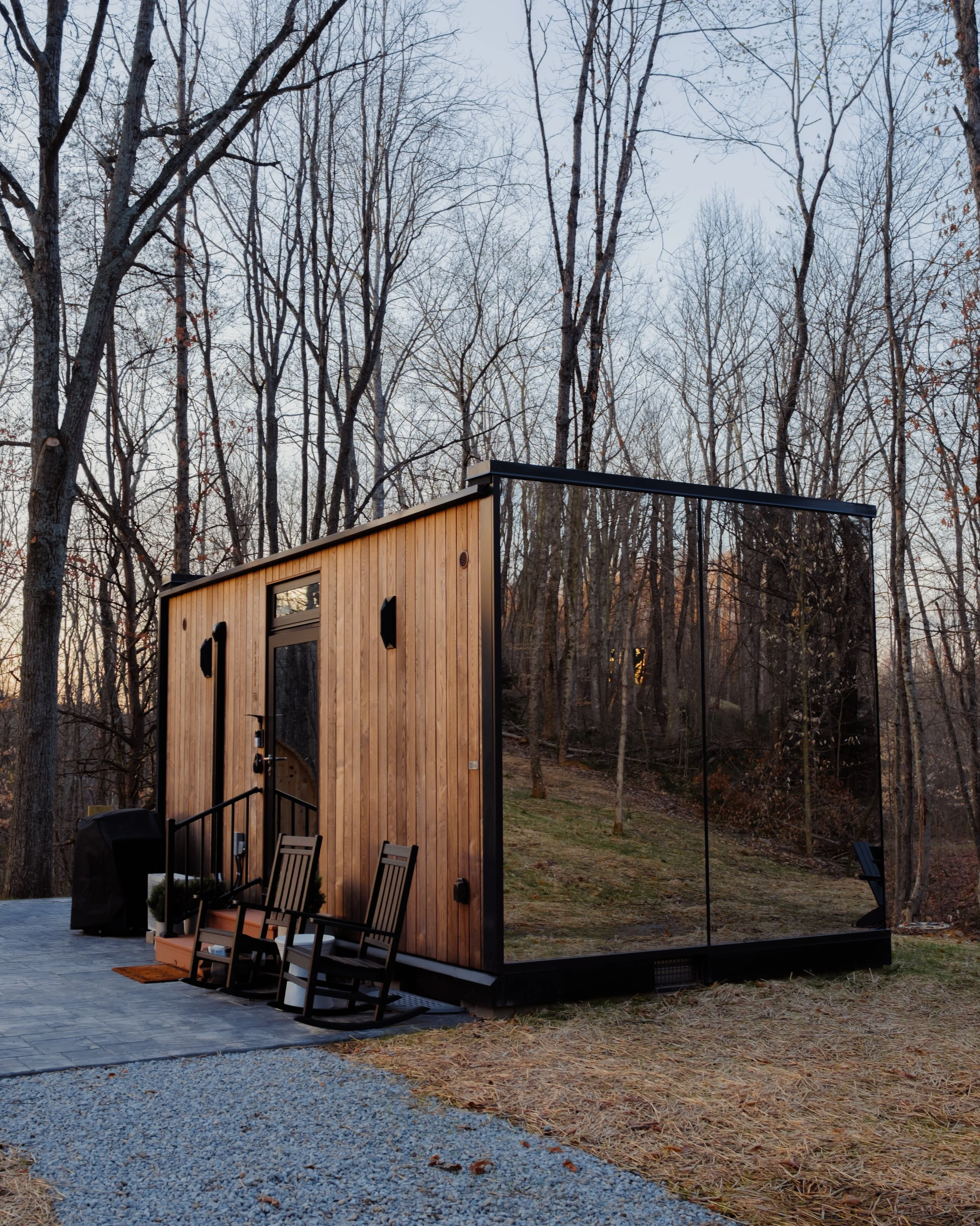 A contemporary wooden tiny house with large black-framed windows in a forested area during late fall or winter, with bare trees and a gravel pathway outside.