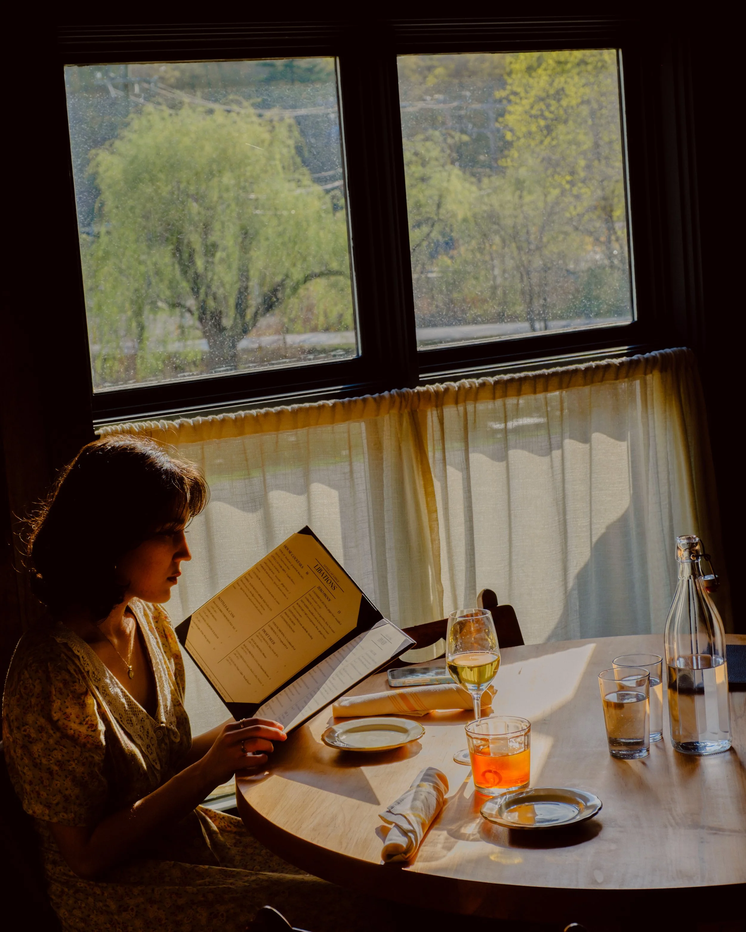 A woman sitting at a dining table reading a menu in front of a large window with curtains, with sunlight illuminating the scene. The table has glasses of water and orange-colored drinks, a bottle, and plates. Outside the window is a view of green tre