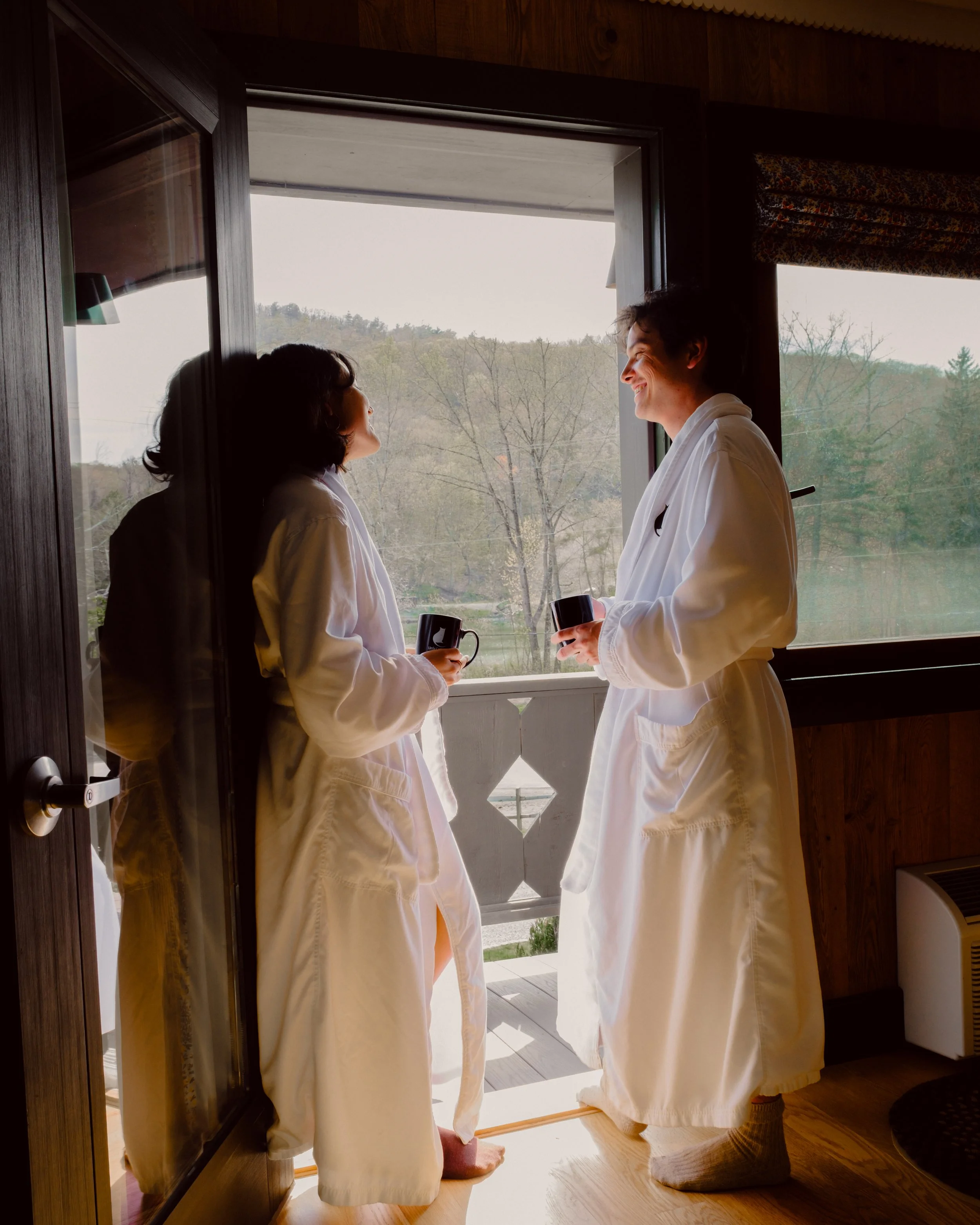 Two people in white robes standing by a door, smiling at each other while holding mugs, with trees visible outside.