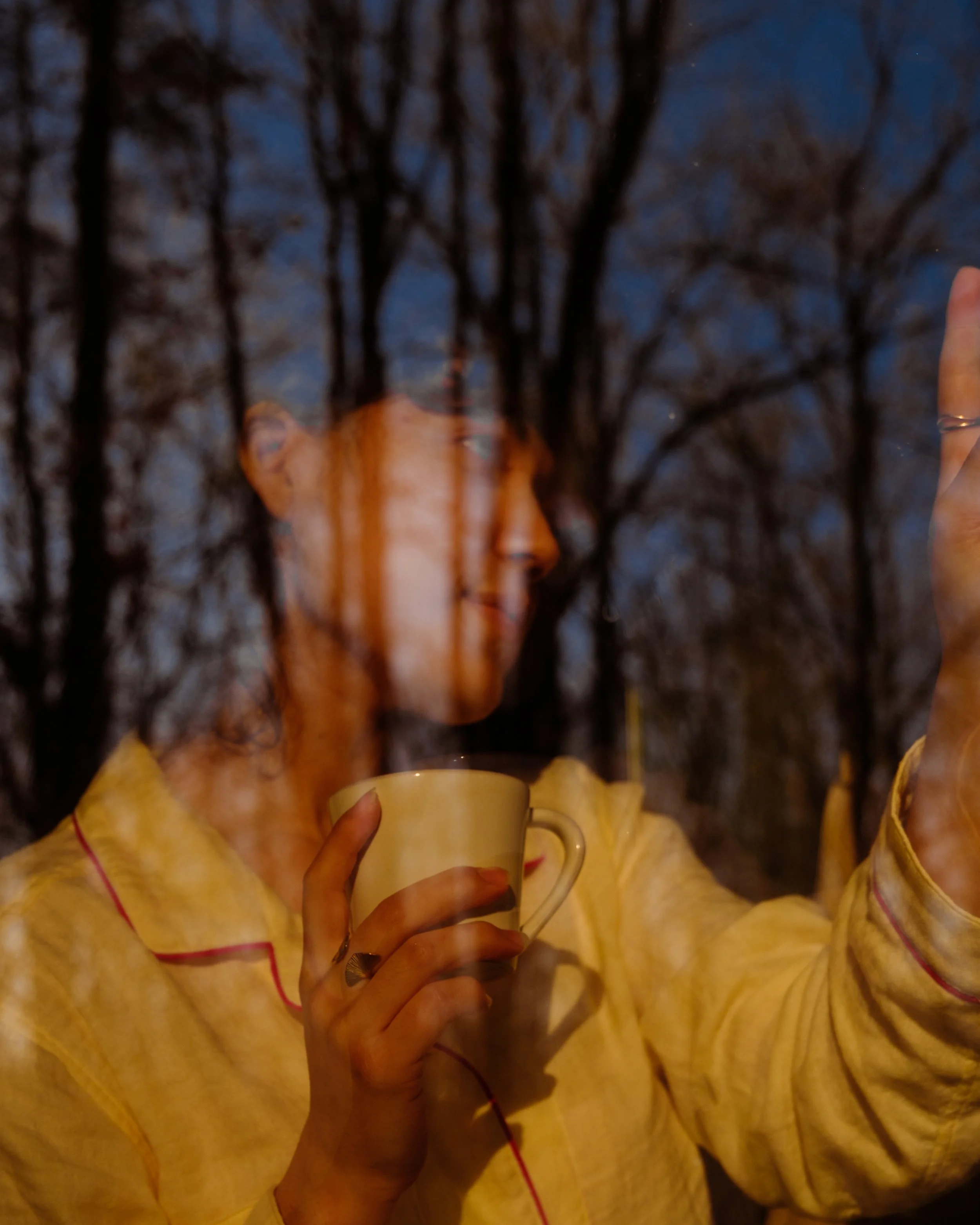 Person holding a beige coffee mug in front of a window at night, with bare tree branches visible outside.