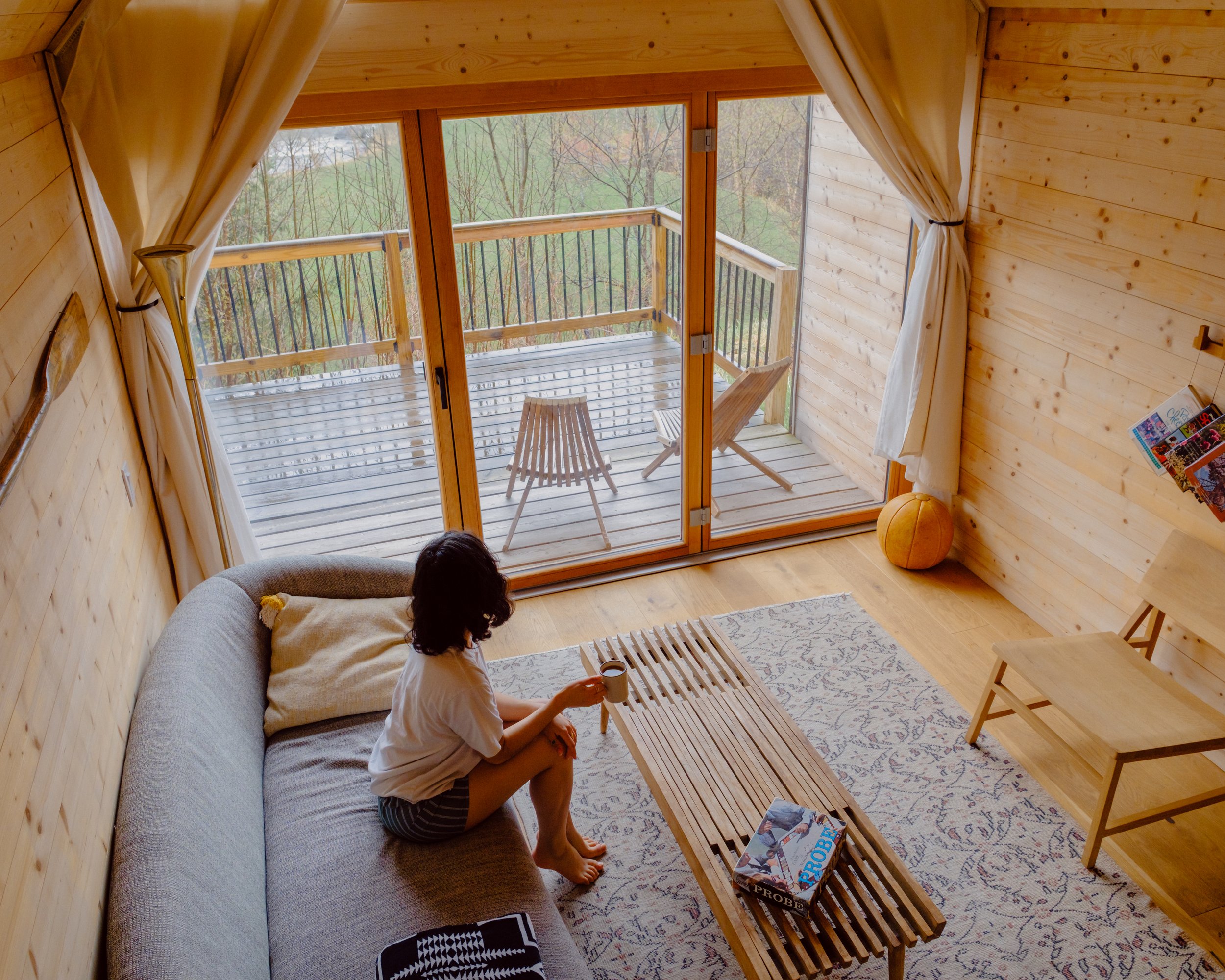A woman sitting on a gray sofa in a wooden cabin, holding a cup. There is a wooden coffee table in front of her, a porch with a wooden railing outside, and trees in the background visible through glass doors. The room has wooden walls and a patterned