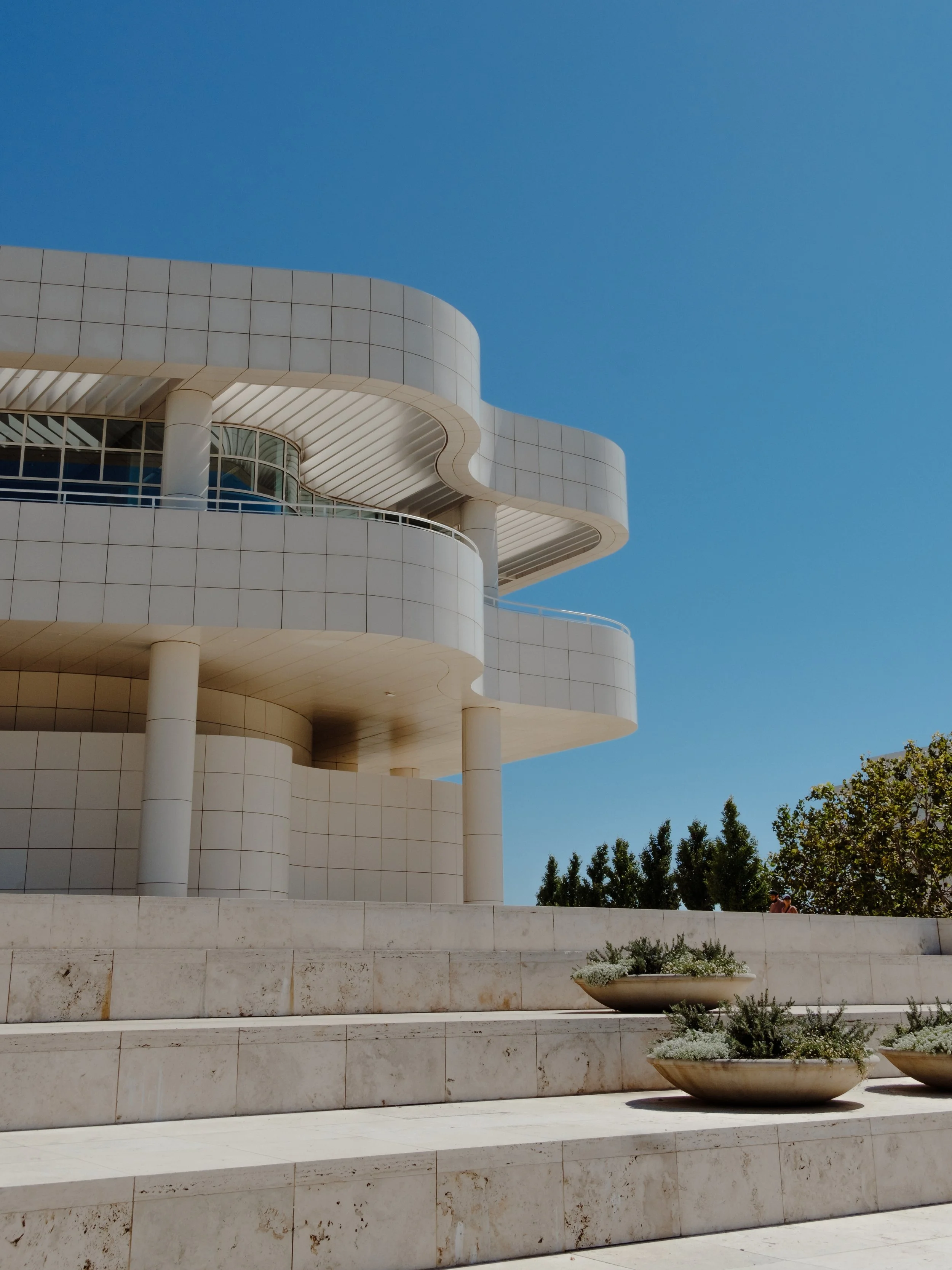 Modern building with curved white facade and glass balconies, set against a clear blue sky, with large planters and some trees at the bottom.