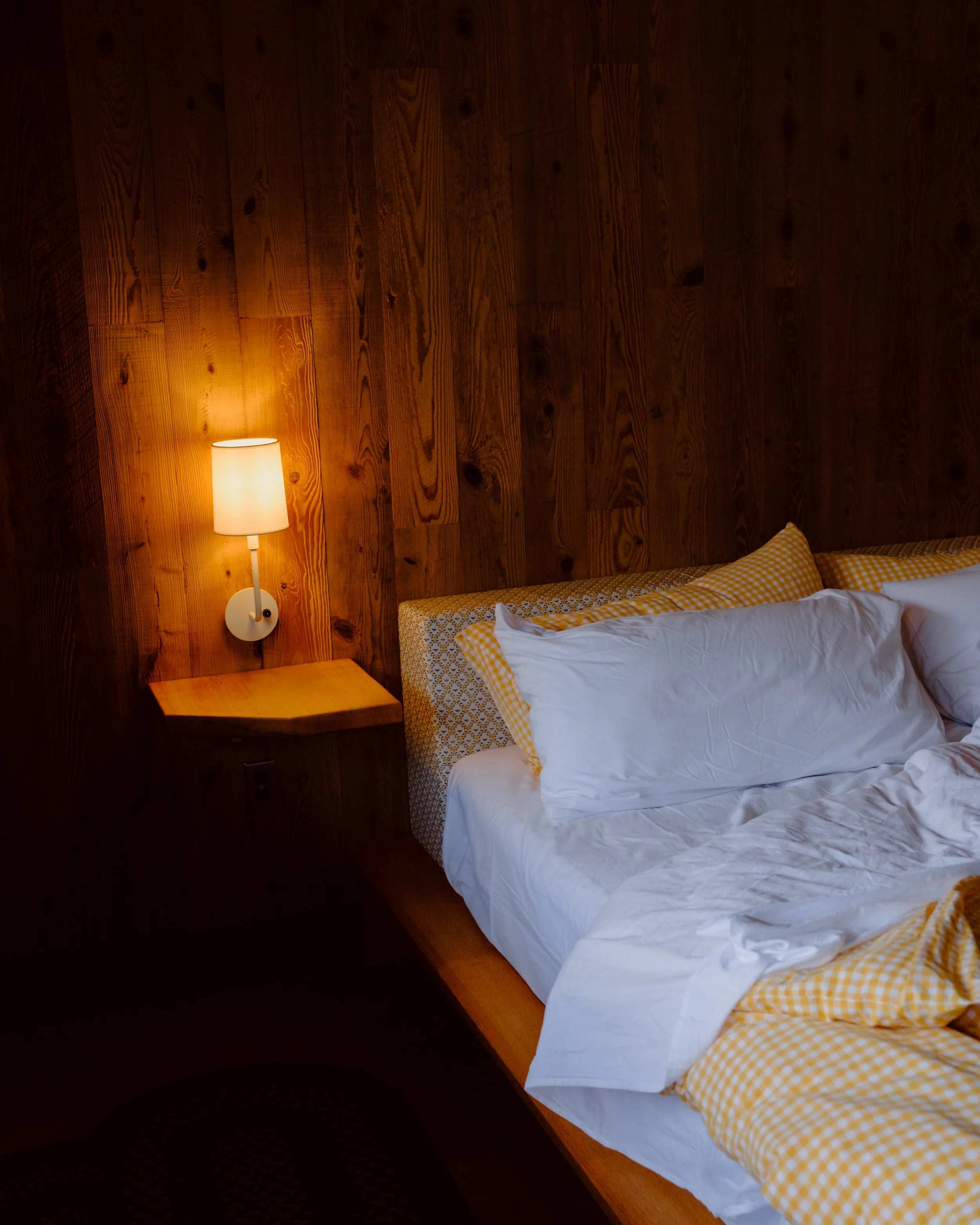 Close-up of a bedroom corner with a wooden wall, a white wall-mounted lamp, and a bed with a yellow and white checkered pillow and white bedding.