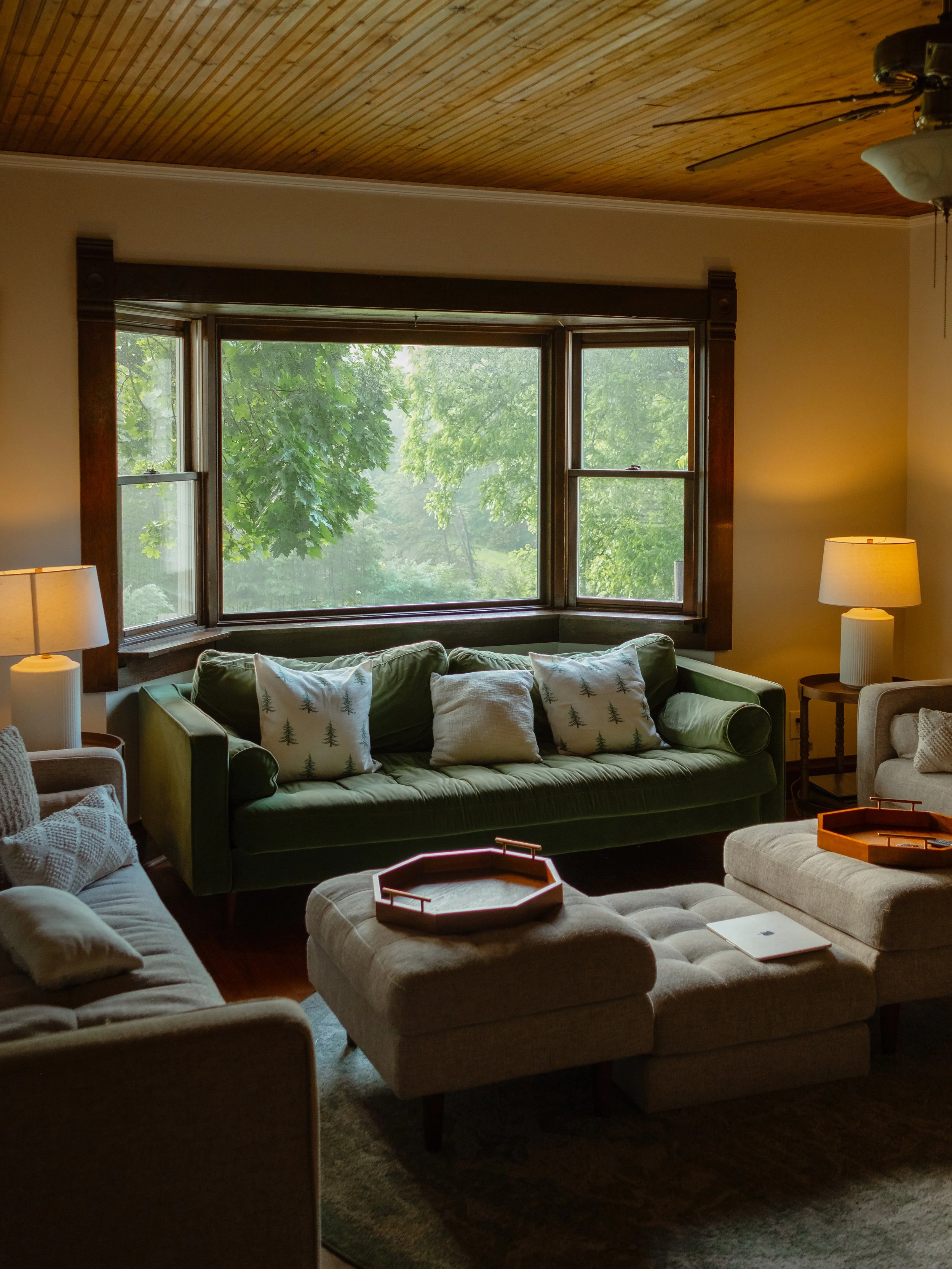 Living room with green sofa, beige armchair, white ottoman, pillows, wooden ceiling, large window with trees outside, side table with lamp, and a tray on the ottoman.