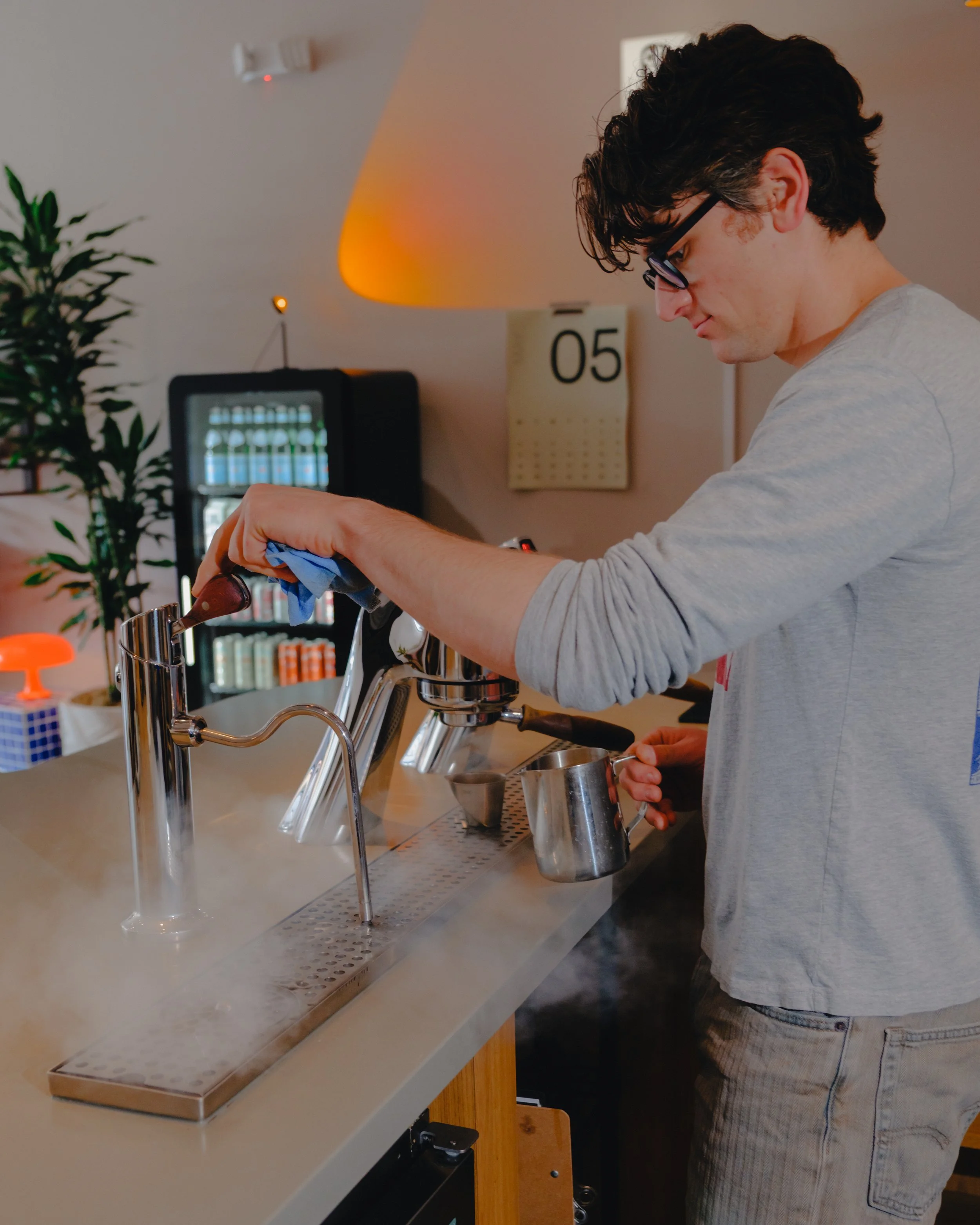 A young man with dark curly hair and glasses is pouring coffee into a cup at a modern kitchen counter. There are shelves with books, a potted plant, a calendar, and a lamp in the background.
