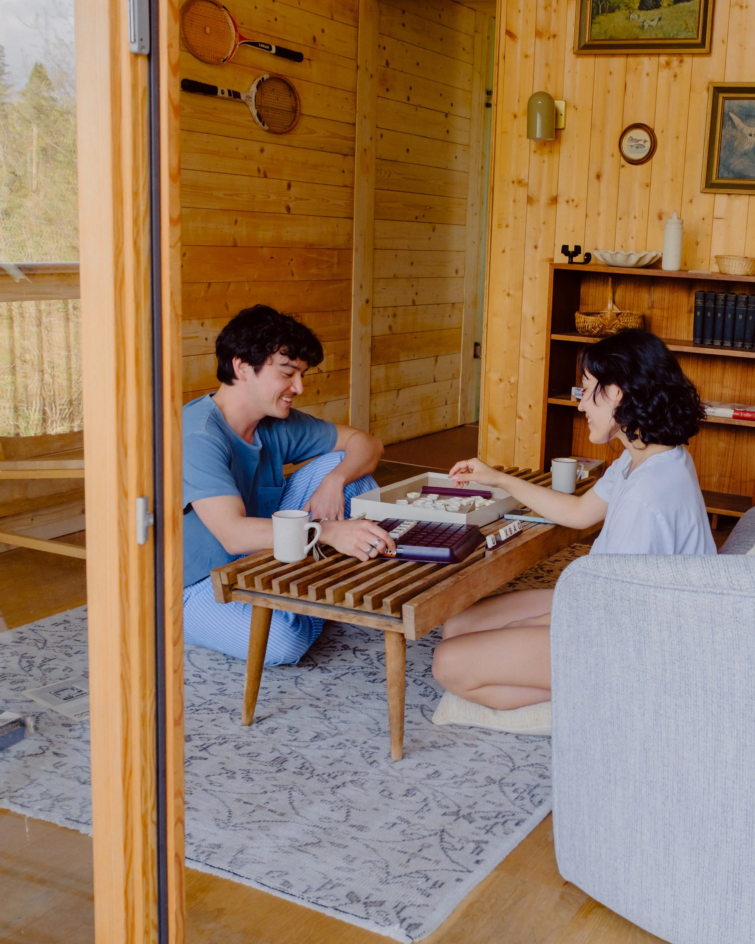 Two people, a man and a woman, are playing a board game at a wooden table in a cozy room with wood-paneled walls. There are mugs on the table, and a bookshelf with books and decorative items is visible in the background. The man is wearing a blue shi