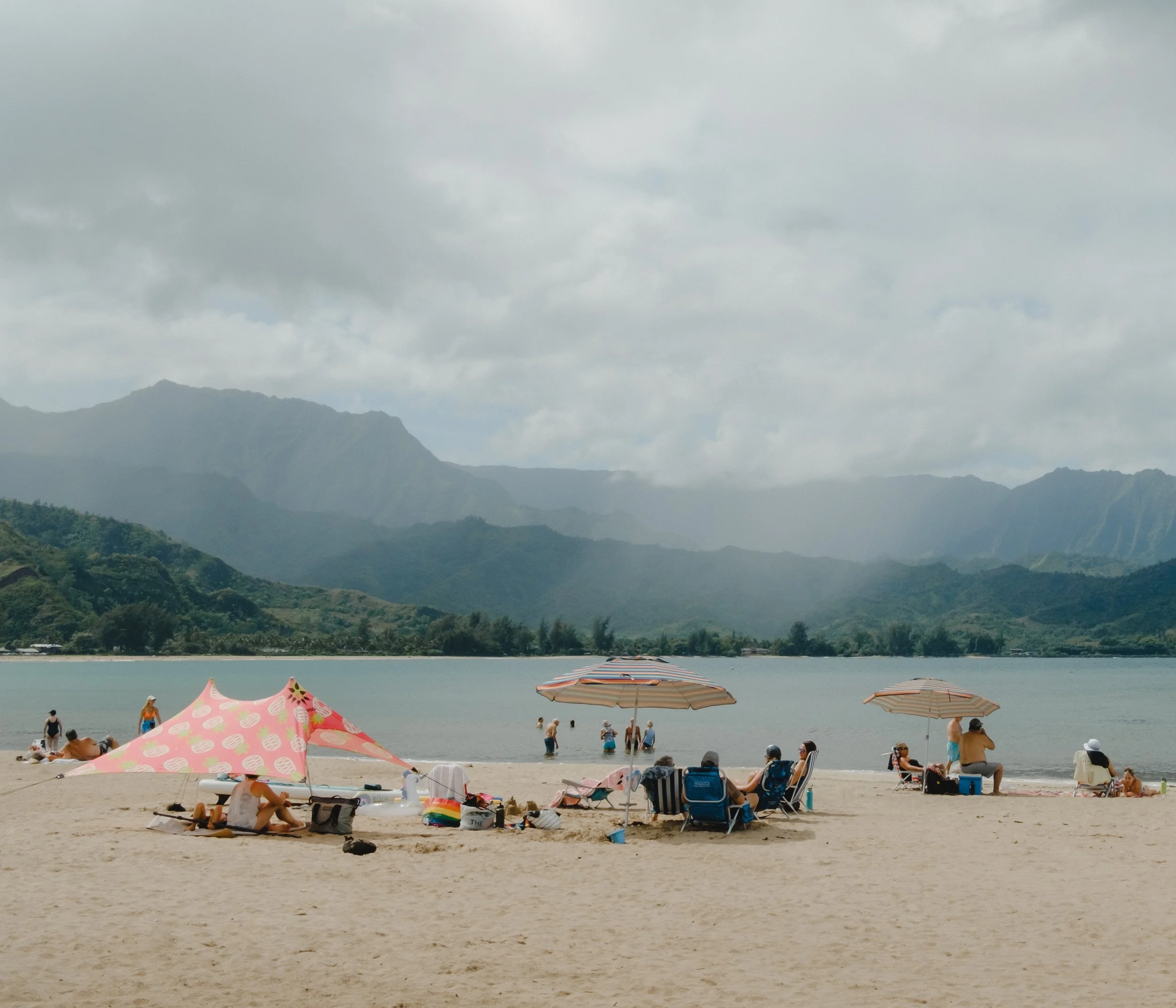 Beach scene with people relaxing under umbrellas and playing in the water, mountainous landscape in the background with overcast sky.
