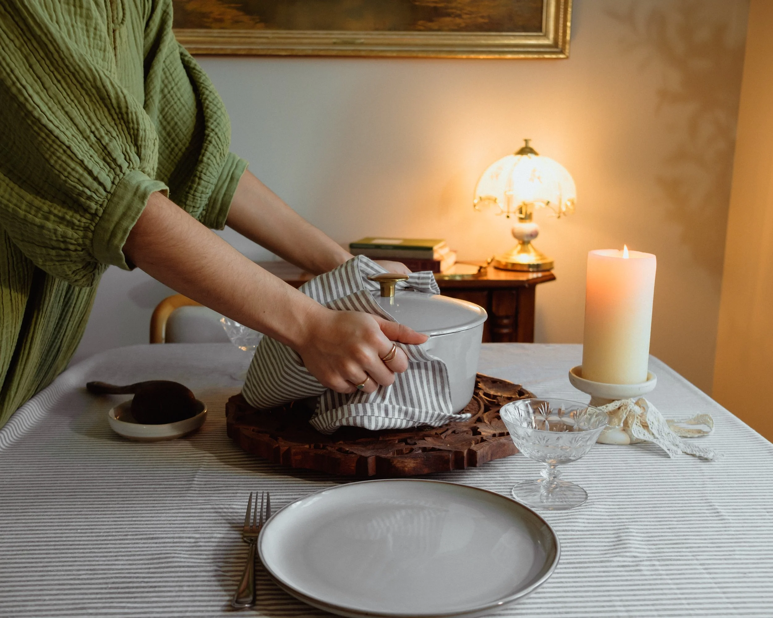 Person wearing a green, textured bathrobe preparing to serve food at a dining table, which is set with a large white candle, a glass bowl of whipped cream, and a plate with chocolate desserts, in a warmly lit room with a framed painting and a table l